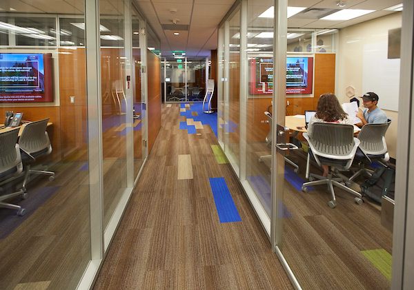 The LL2 Consultation Space. Two rooms are featured on either side of the hallway. In the righthand room, two people are meeting around a table. 