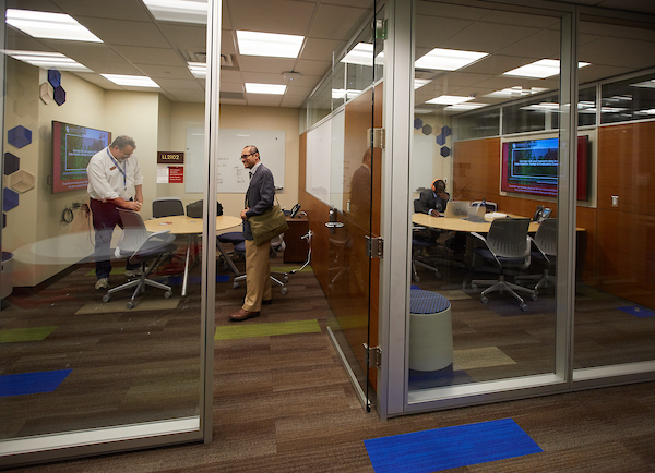 Two meeting rooms side-by-side. One features two people standing and talking with one another. In the other room is a student working on their laptop at a table. 