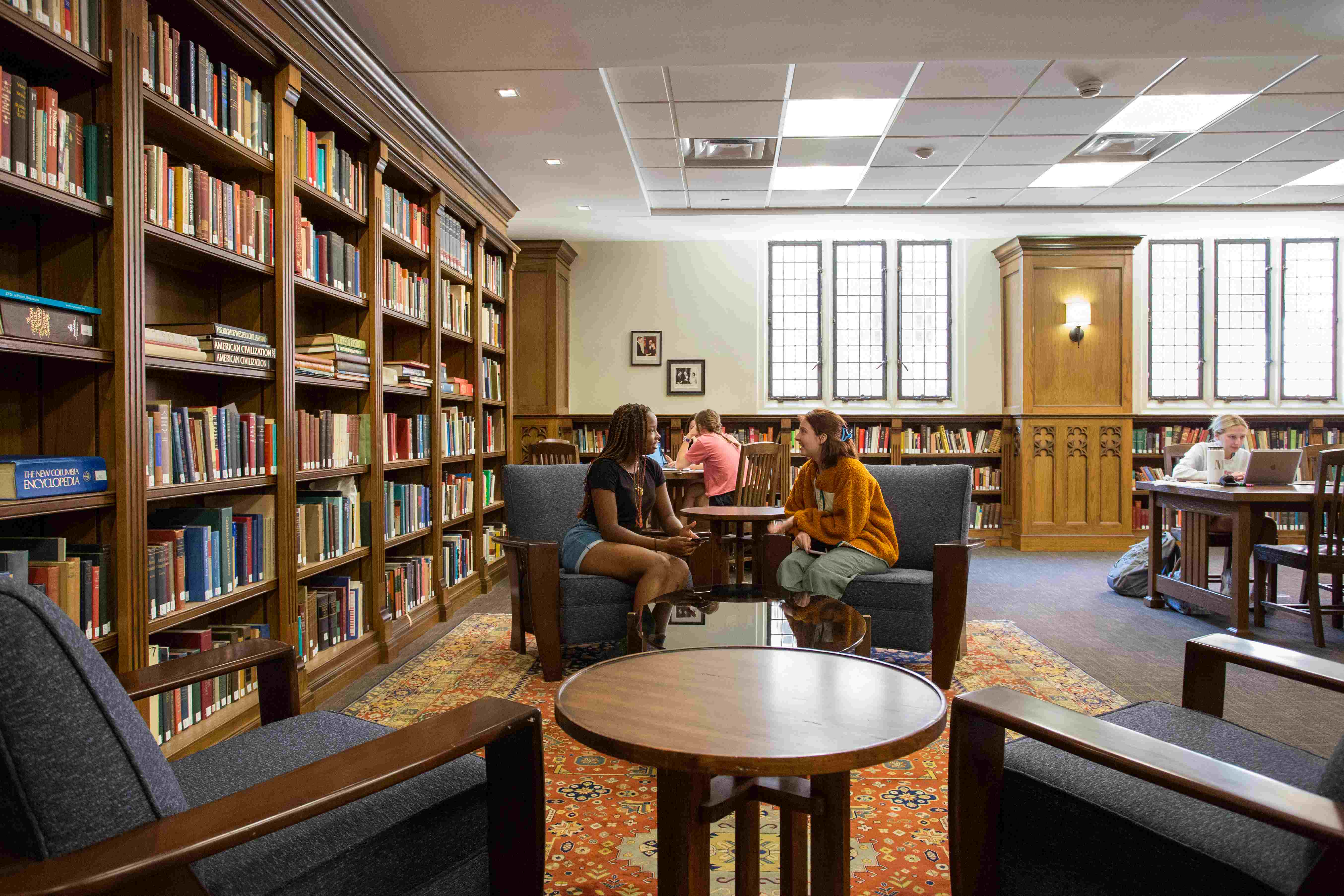 This view of the Boorstin Collection room shows multiple bookshelves and a seating area. Two students are sitting in chairs next to one another and are talking over a side table. 