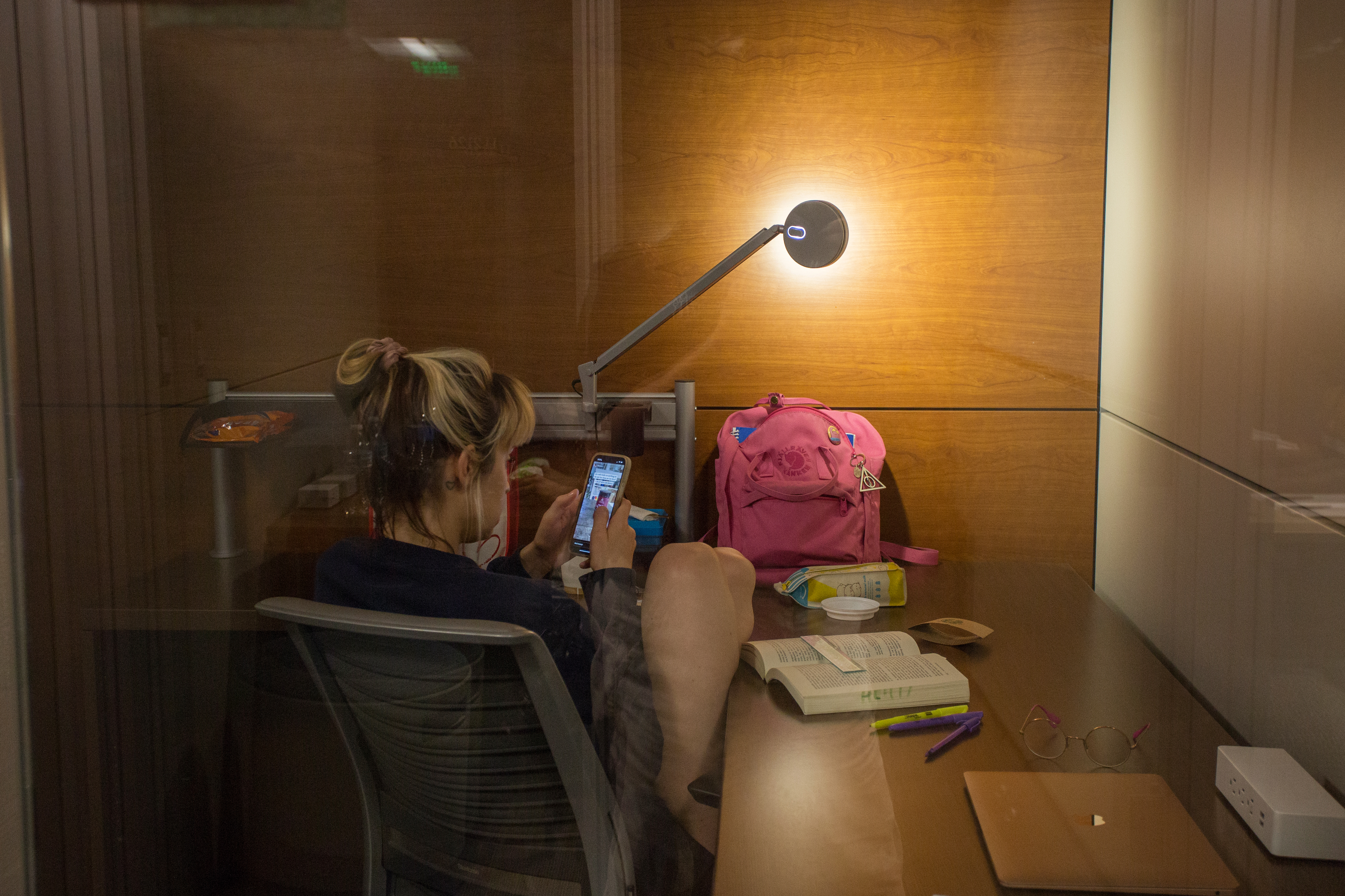 Student sits at a corner desk looking at their phone. On the desk is an open book, a bag, a laptop, glasses, and highlighters. 