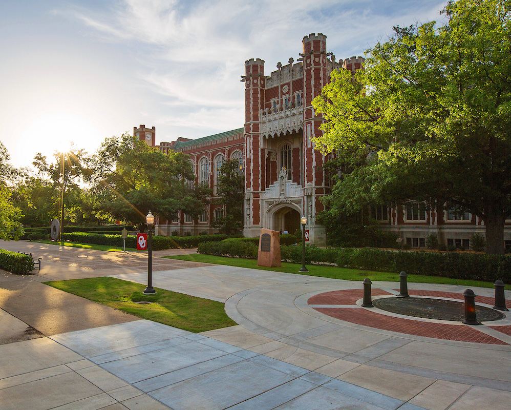 An exterior, angle shot highlights the ornate architecture of Bizzell Memorial Library's South Entrance. It is late afternoon and the sun is beginning to set behind the beautiful green trees and bushes on either side of the entrance.
