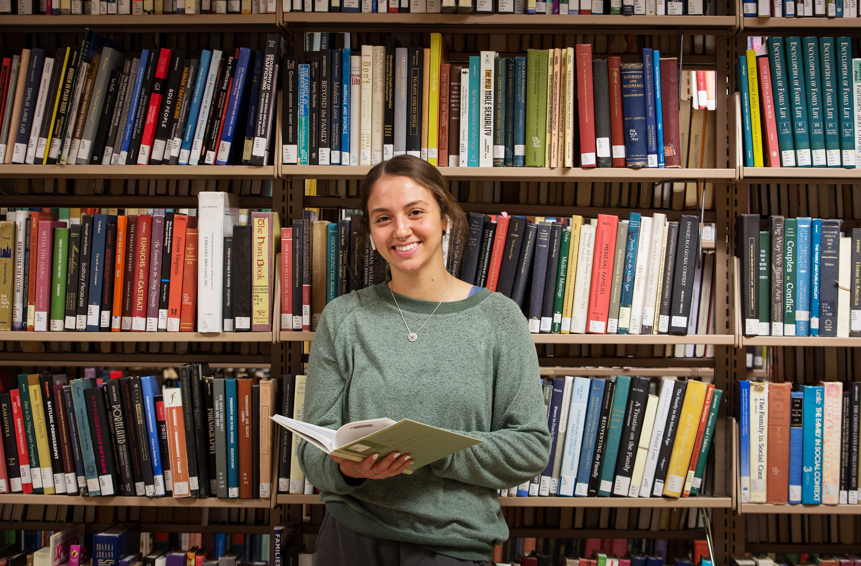Happy female student is standing in front of a shelf of books in the stacks and smiling at the camera