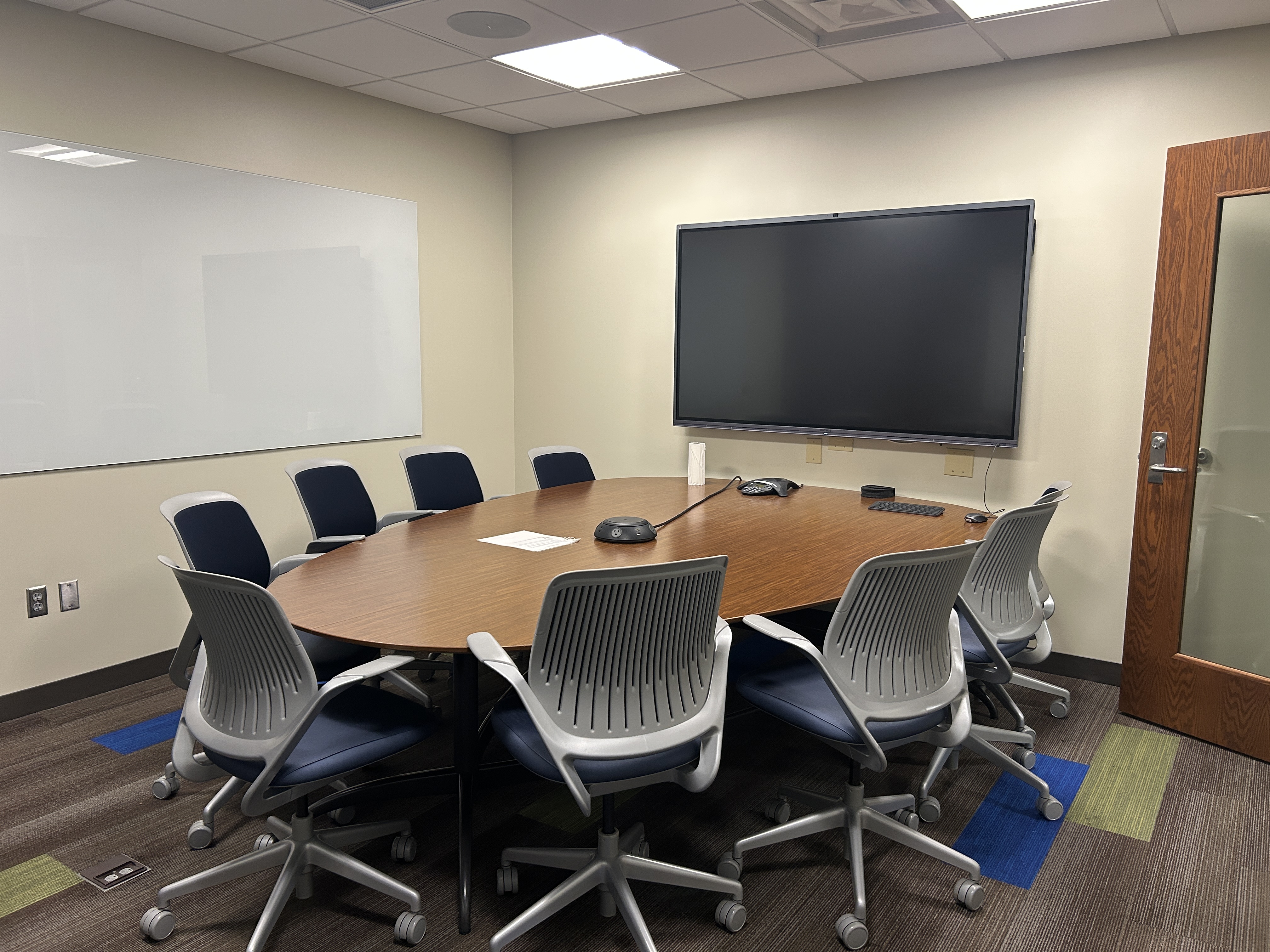 A room with a whiteboard on one wall and a large television screen on the next. In front of the television is a table with eight chairs around it. 