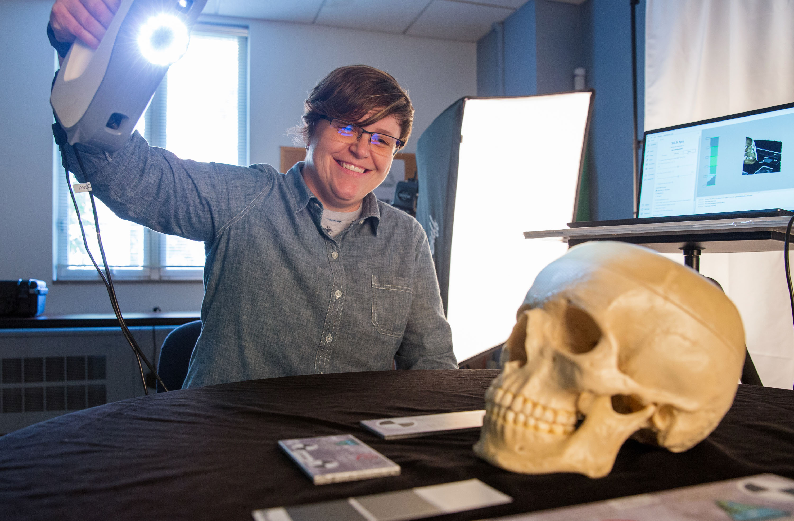 Lab staff member is holding a scanning device above a human skull to scan the skull image to a laptop behind her.