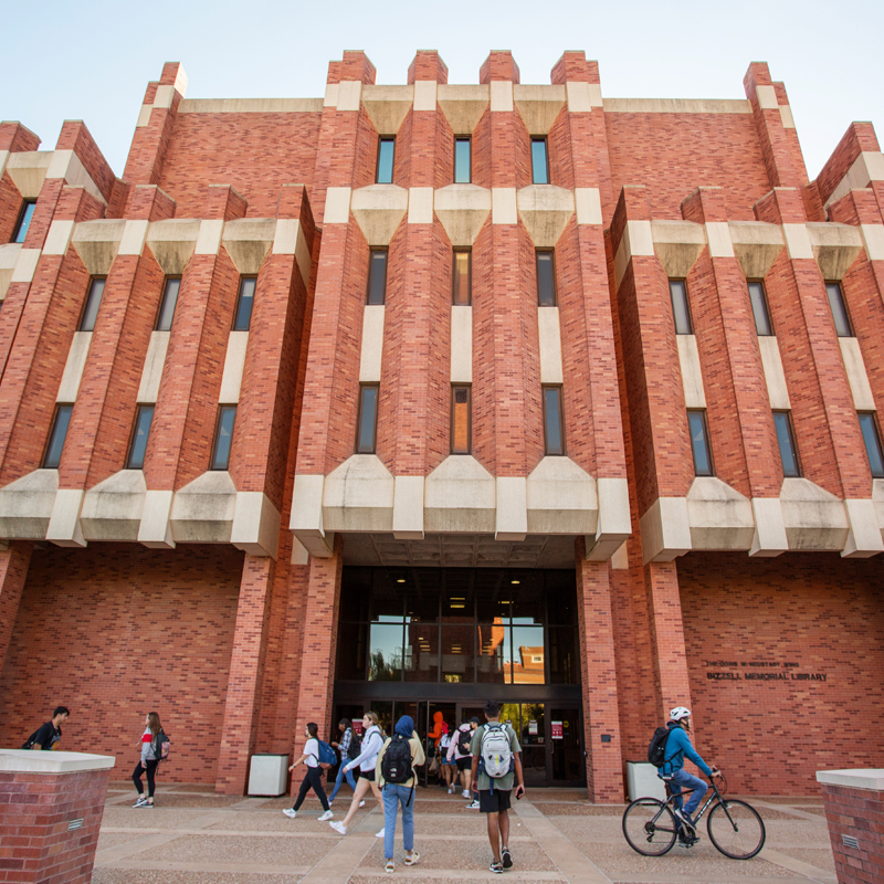 Bizzell Memorial Library west entrance showing red brick facade with students in front walking and biking.