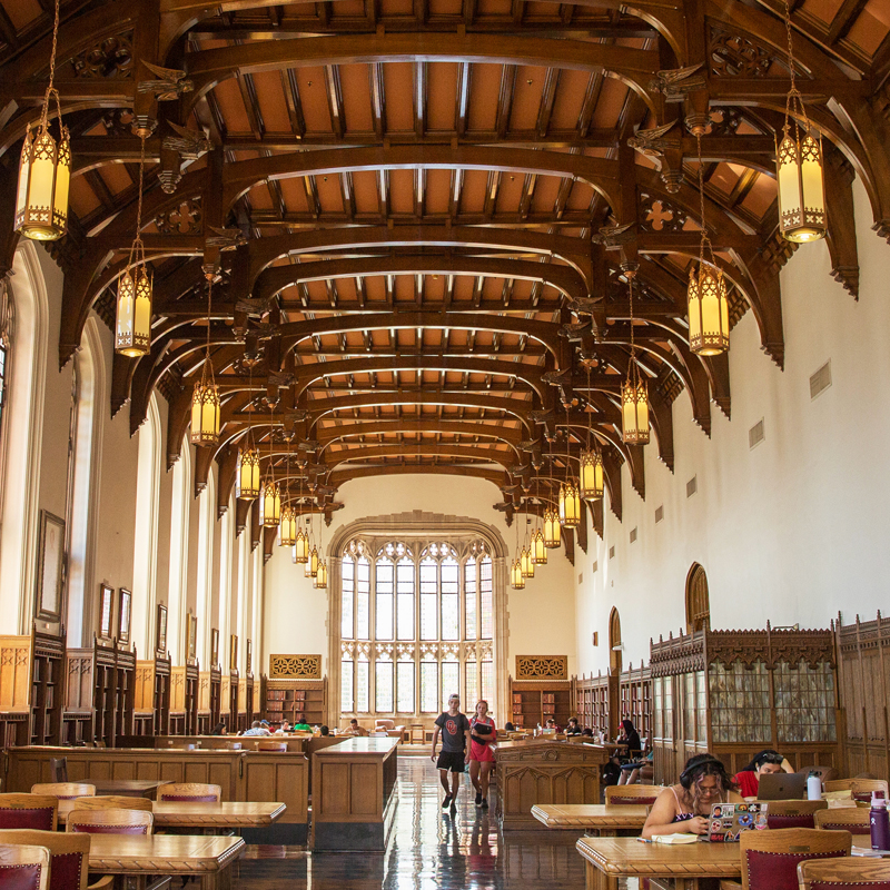 Bizzell Memorial Library's Great Reading Room features ornate architectural decorative elements with high, dark wood ceilings and tall, multipane windows. The walls are lined with carved, dark wood bookcases that hold theses and dissertations of OU graduates. Students study at various tables through the room and a pair of students walk towards the camera in the center aisle of the room between the tables.