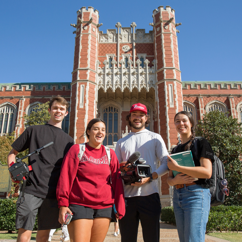 In the background, Bizzell Memorial Library's south entrance has ornate architecture and red brick facade. A group of four smiling stand in front of the building with backpacks on their shoulders while they hold books and camera equipment