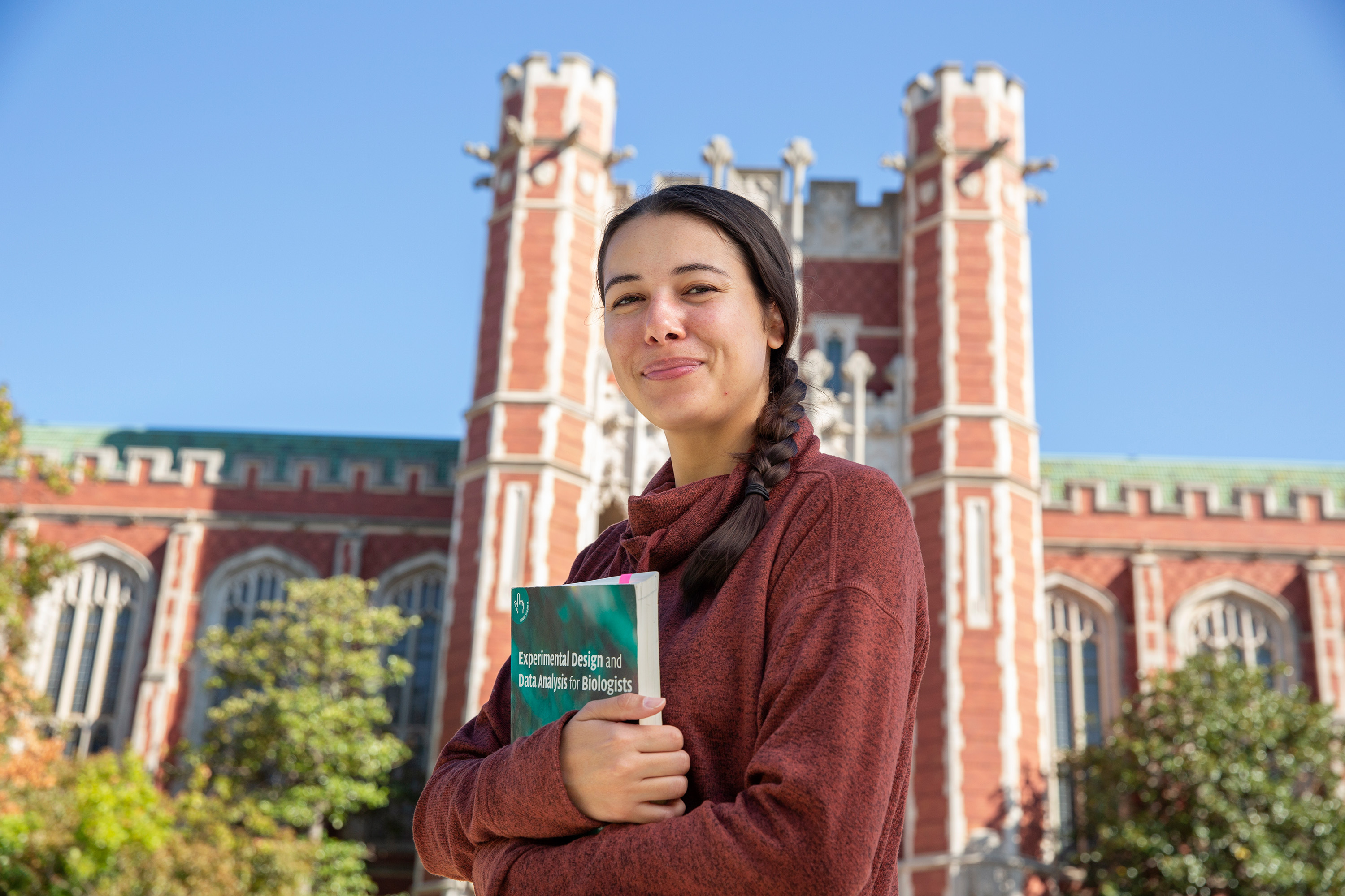 Brunette, female student is holding a book and smiling at the camera while standing in front of Bizzell Memorial Library on a sunny day with blue skies in the background.