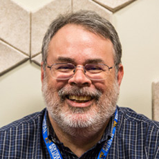 Mark Laufersweiler is wearing a blue plaid shirt and wire rimmed eyeglasses and smiles for the camera.