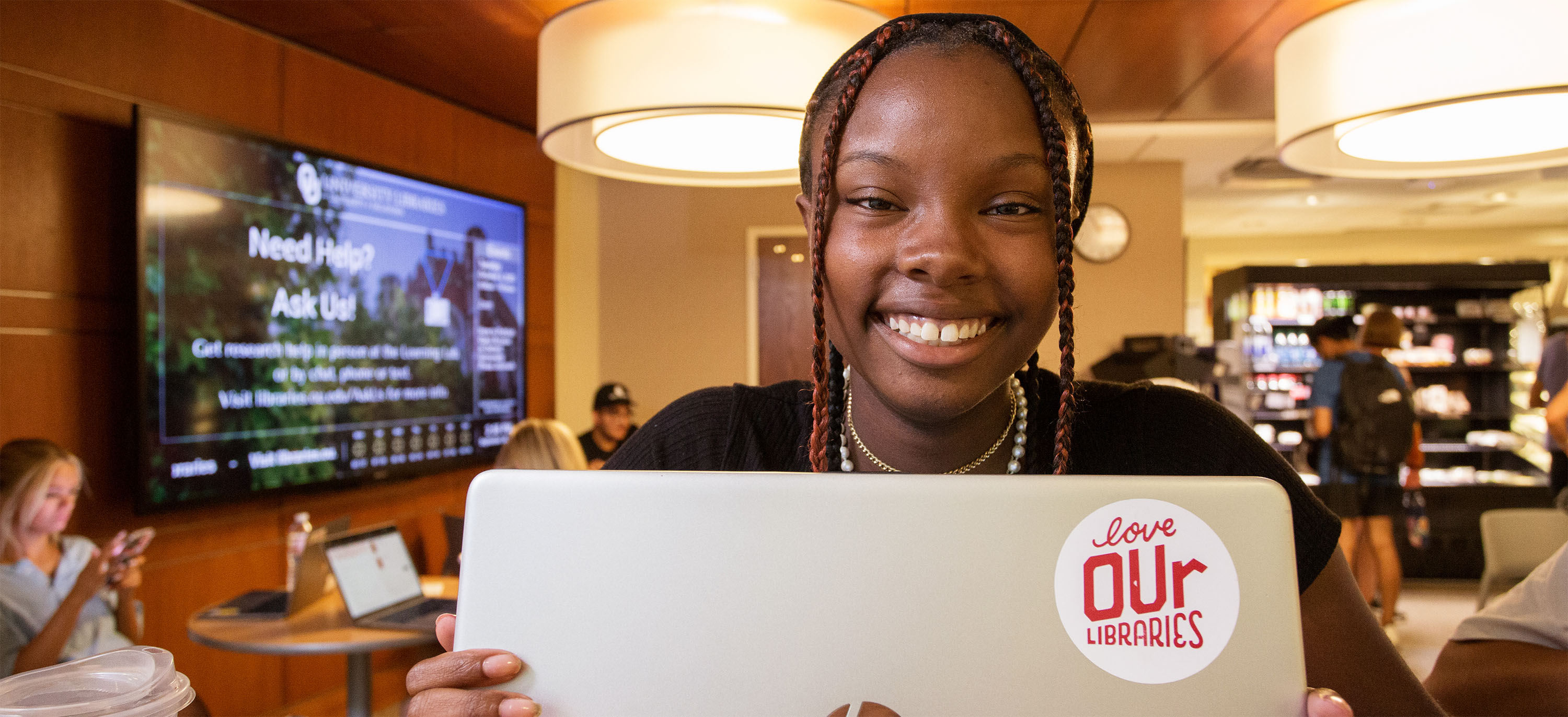 Female student is sitting at a table, facing the camera and smiling with her laptop in front of her. On the back of her laptop is a "love OUr Libraries" sticker. She is sitting in the Bookmark Cafe and there is a energetic scene behind with students sitting at tables and getting drinks and snacks in the Cafe.
