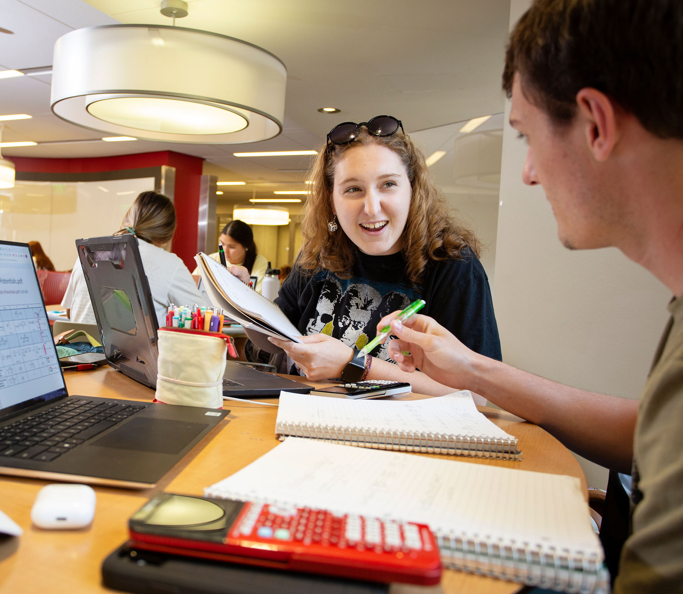 A male student and a female student are collaborating in the Helmerich Collaborative Learrning Center. They are talking and smiling with laptops, notepads, pens, and calculators in front of them at the table they are sitting at.