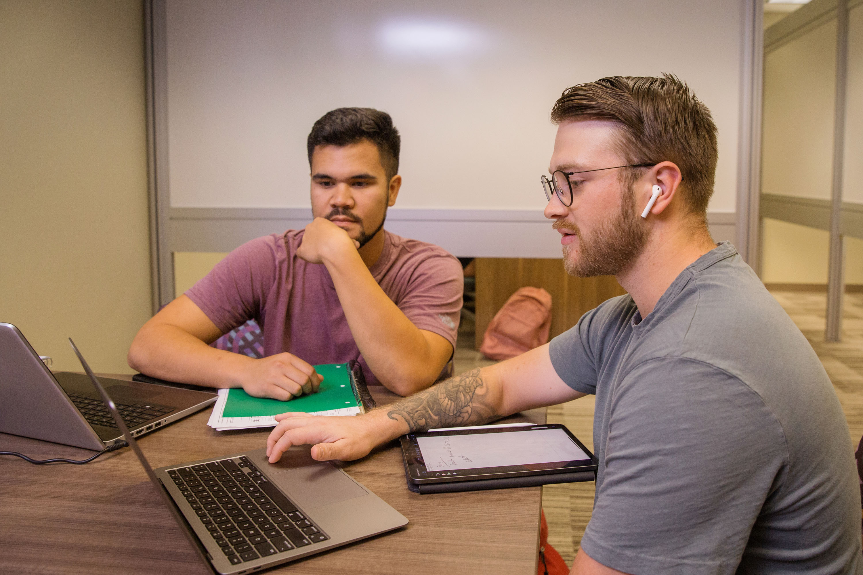 Two male students sit at a round work table collaborating as they look at their laptops on the table in front of them.