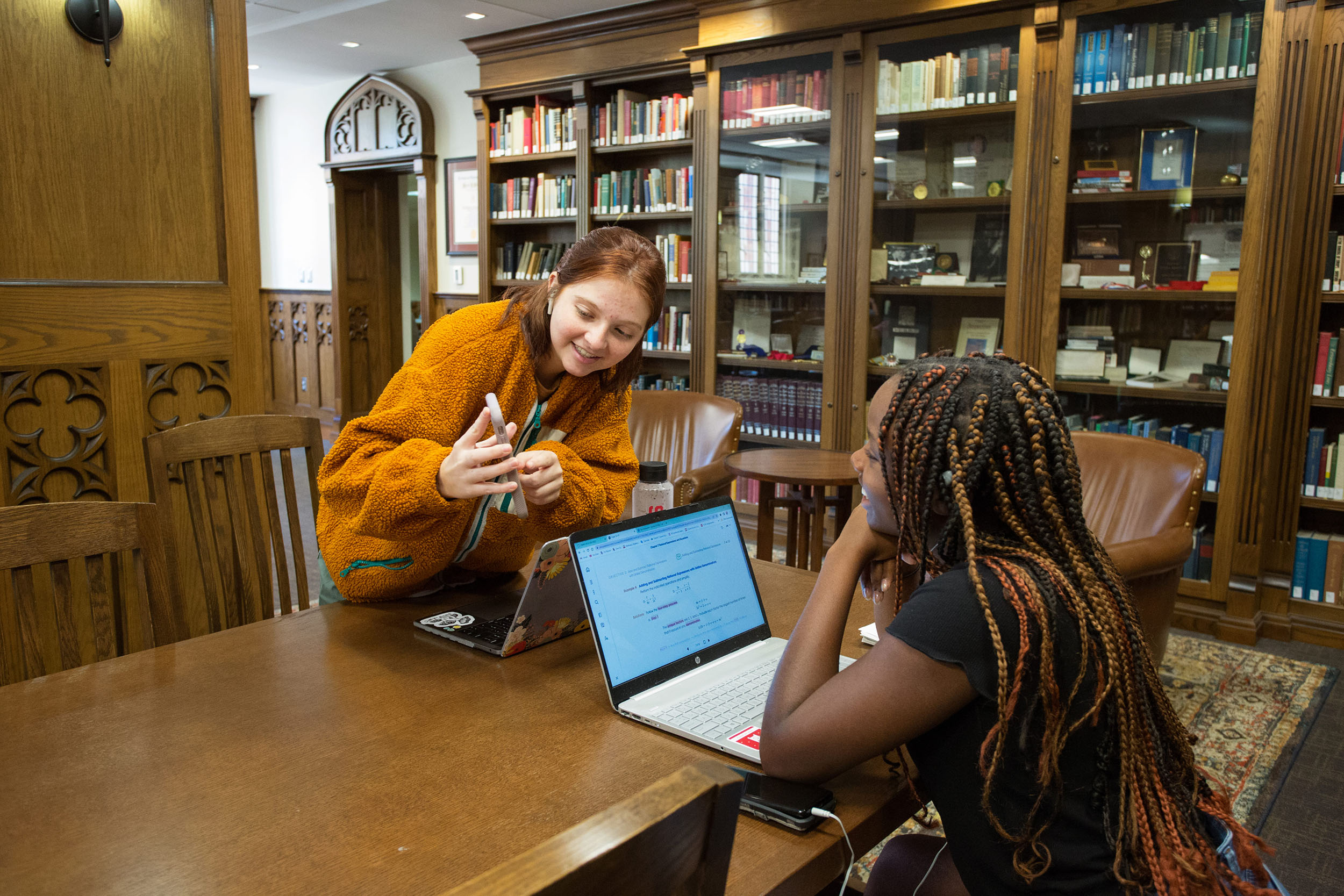 Two women in a library are sitting across from each other at a long, narrow table with their laptops in front of them, smiling. One leans over the table to show her friend something on her phone. In the background, wooden shelves with books line the walls.