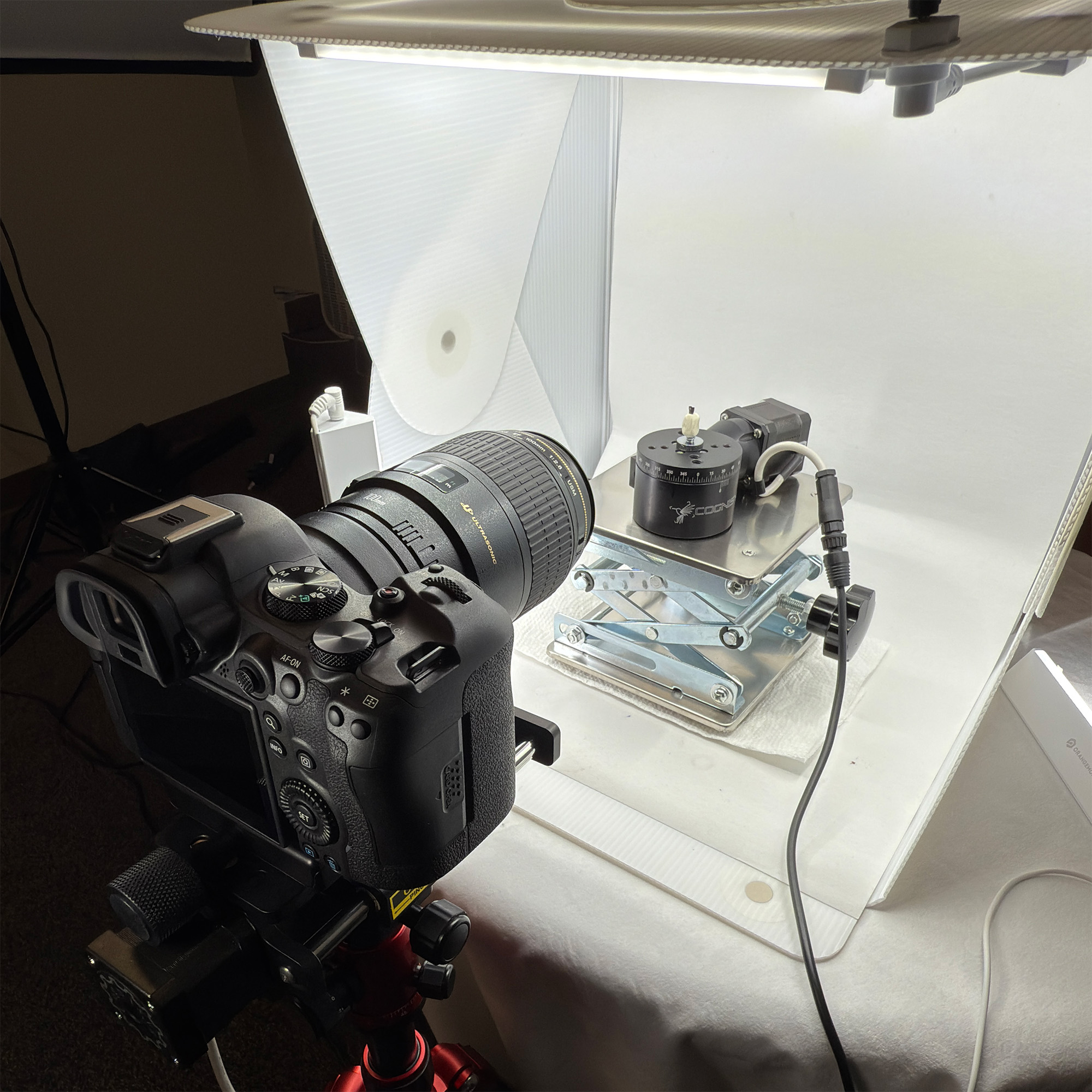 A DSLR camera is set up on a tripod, facing a mechanical device with wires, in a bright, white studio setting. The scene conveys a professional and technical focus.