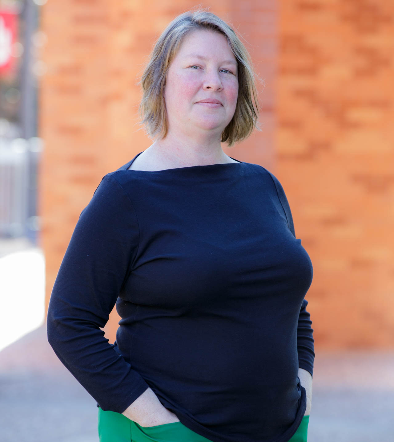 Amanda Schilling is wearing a long sleeve navy blue shirt and stands in front of a red brick building, tucking her hands into the front pockets of her green pants.