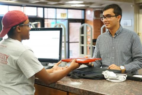 A library employee is standing behind the circulation desk with a computer on the left side of the desk. He is assisting a library patron who strands across the desk and is being handed a piece of technical equipment to borrow.