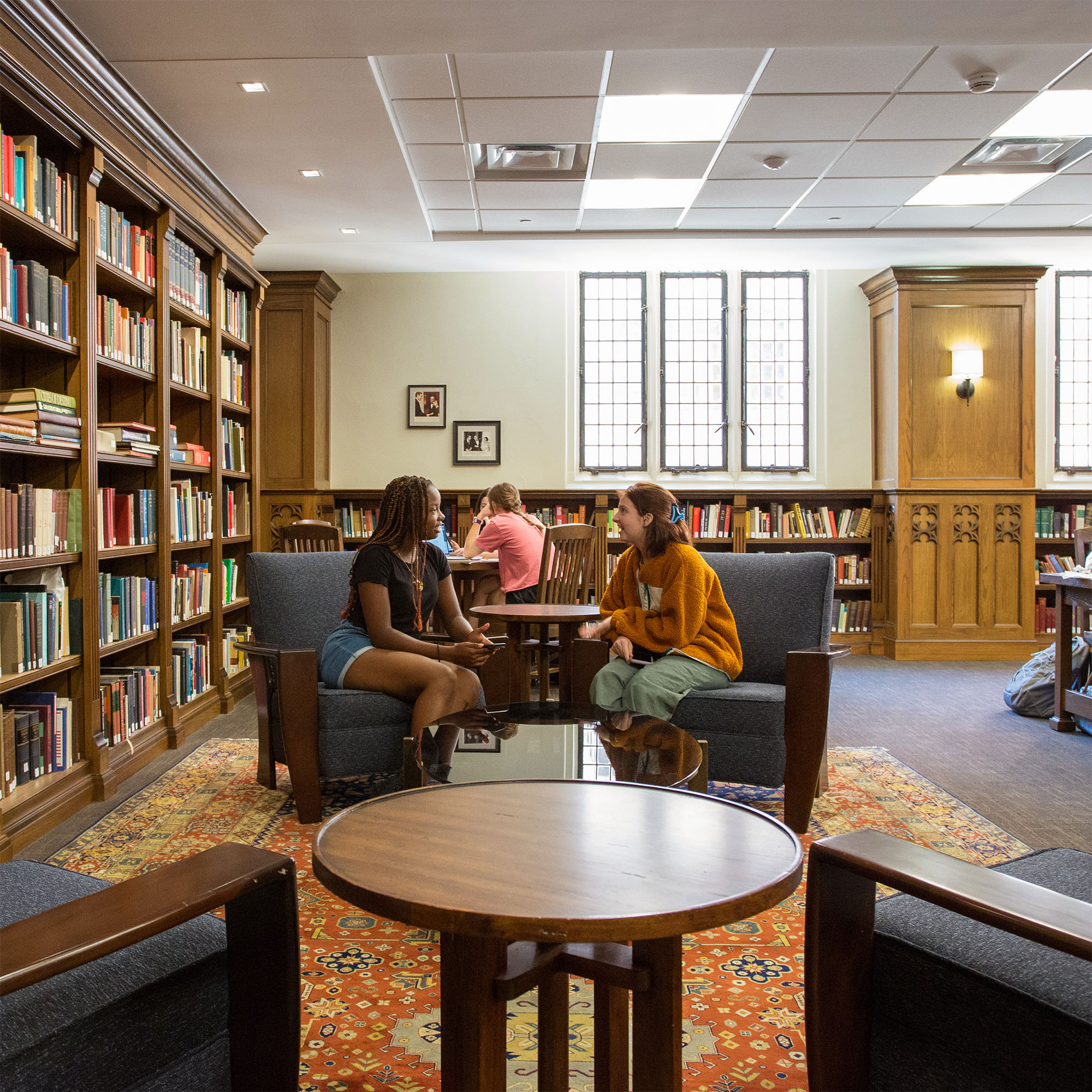 Two people sit on armchairs and chat in a cozy library with dark, wooden bookshelves and tall, narrow windows in the background. 