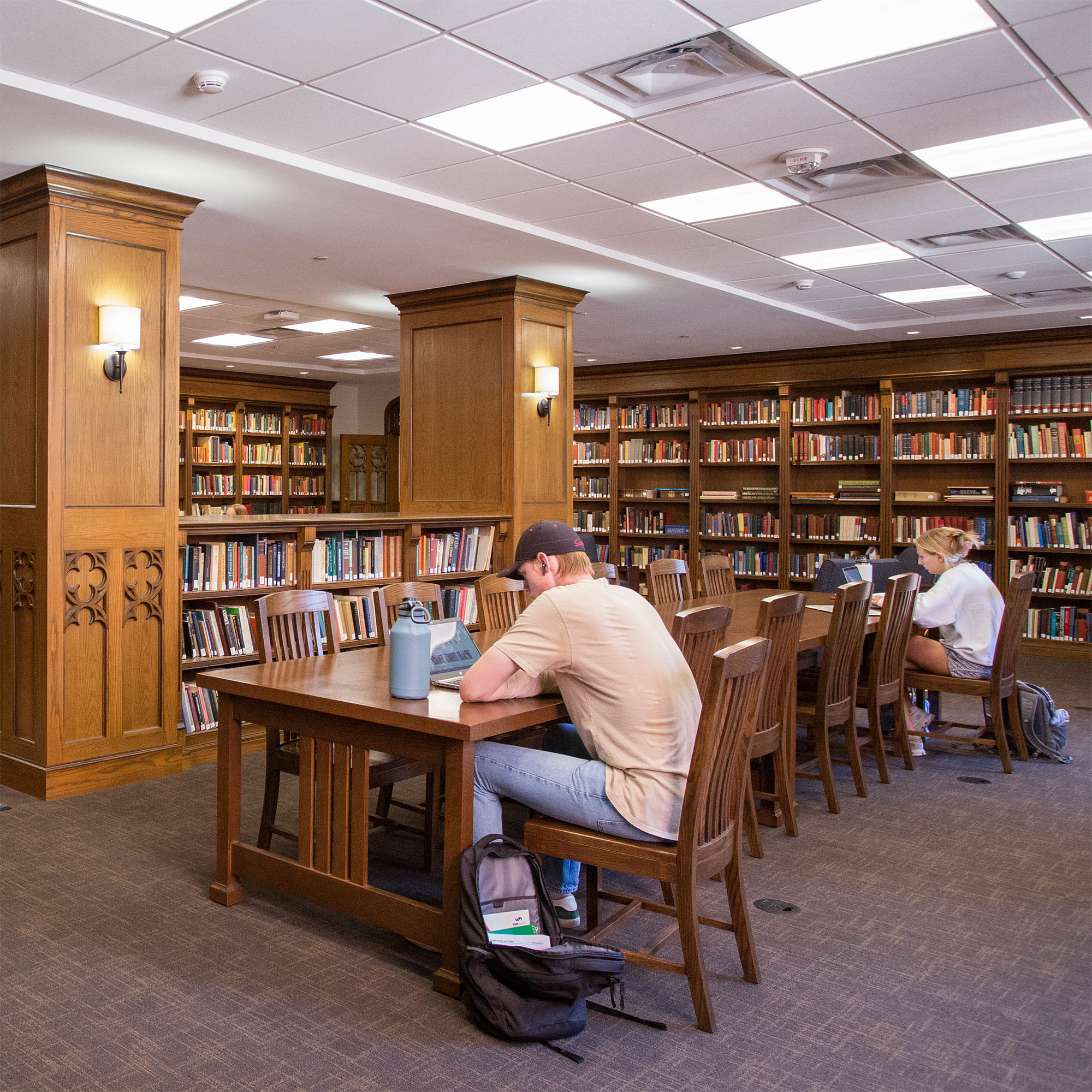 A quiet library filled with dark, wooden bookshelves where people sit at the tables studying on laptops and books.