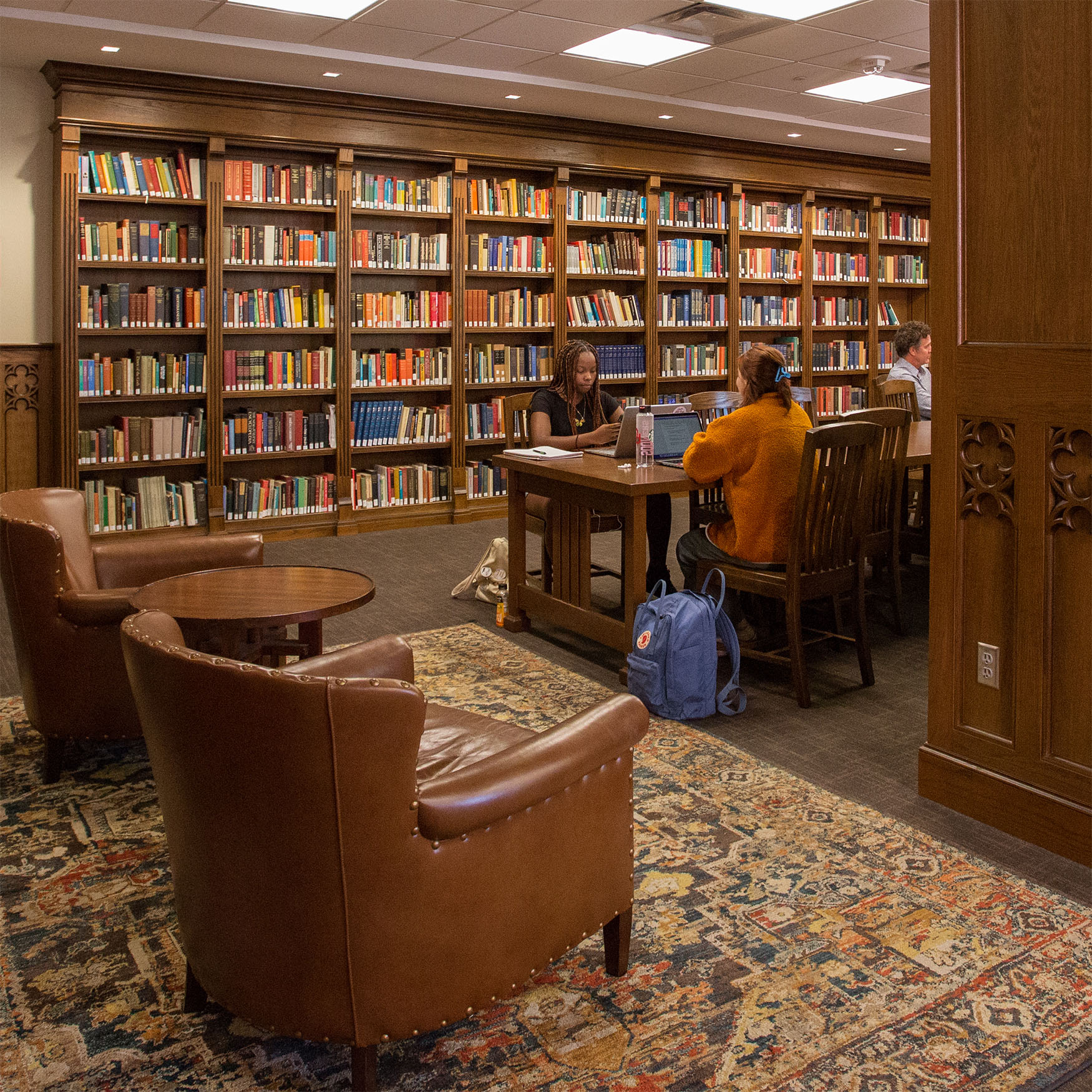 A cozy library with wooden bookshelves filled with colorful books. Three people study at a wooden table, creating a focused and serene atmosphere. Two leather chairs and a patterned rug add warmth.