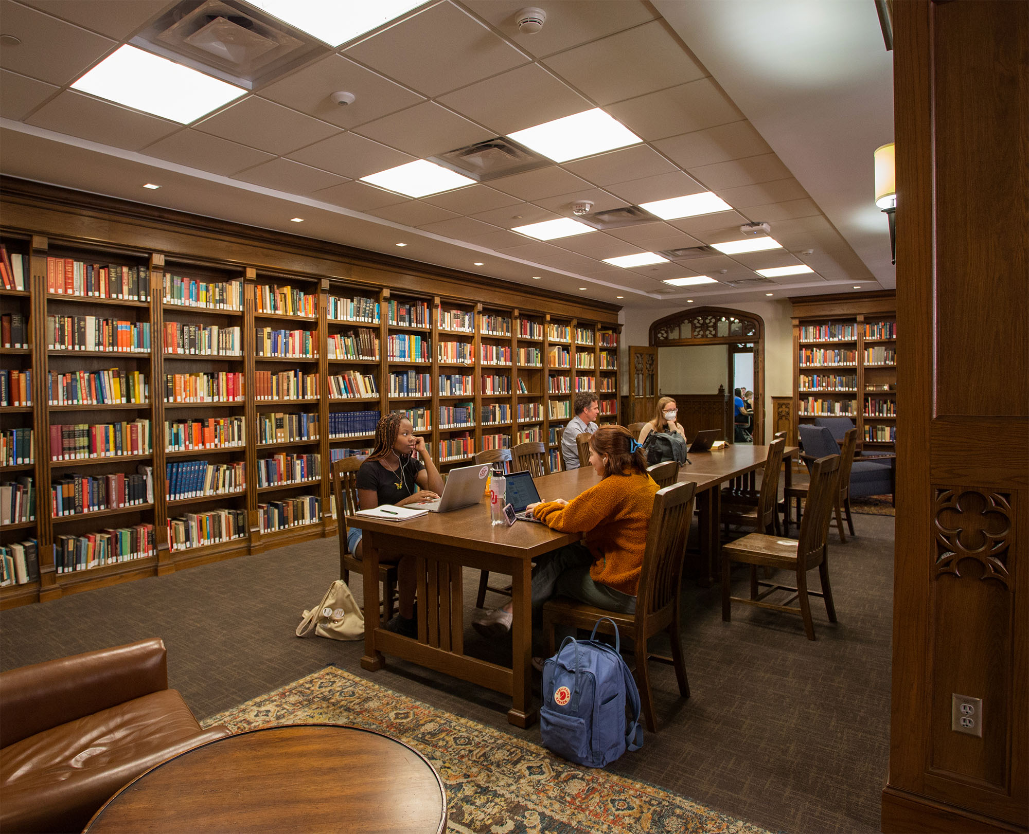 A library room with dark, wooden bookshelves filled with colorful books. Four people sit at a long table, studying on laptops. A dark brown, upholstered chair with a small round table is in the foreground.