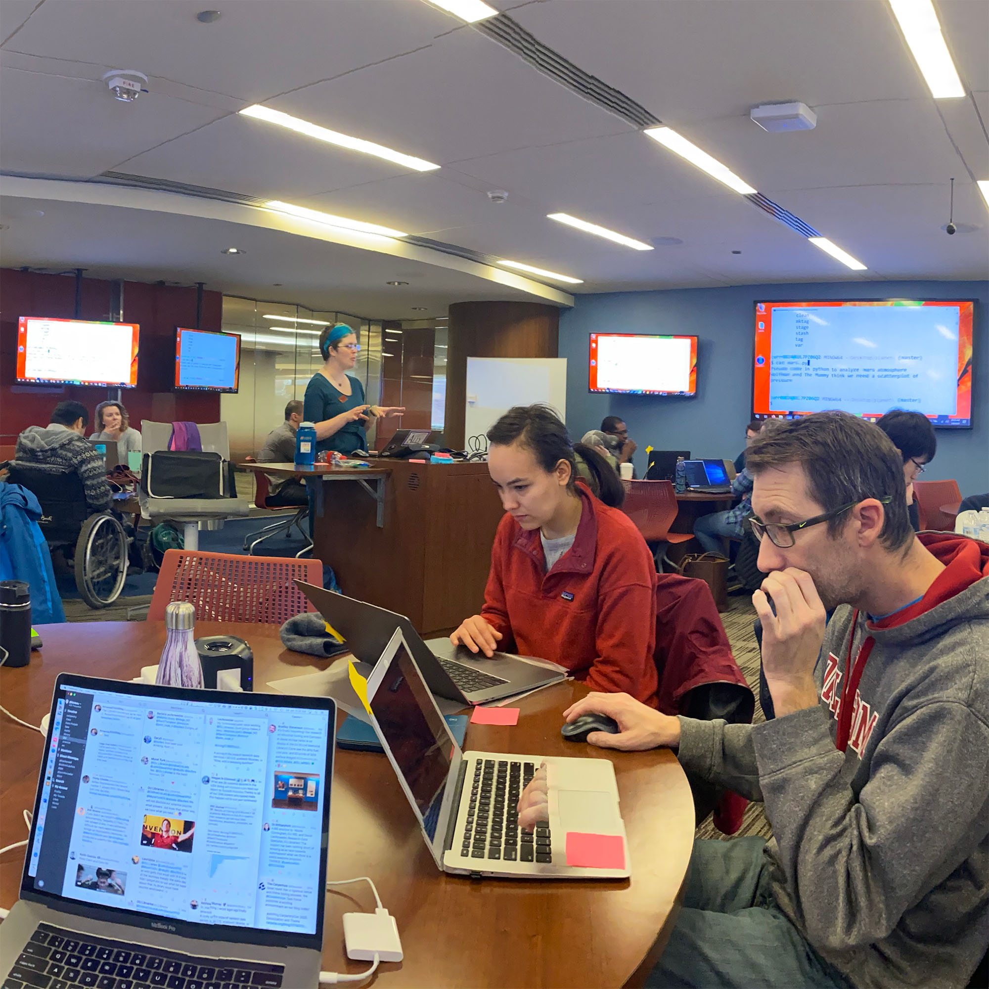 Students sit at round tables working on laptops in a workshop session while the instructor stands at a presentation desk at the center front of the room with her laptop on the desk. Large digital displays are on the walls around the room so students can follow what the instructor presents on her laptop to the class.
