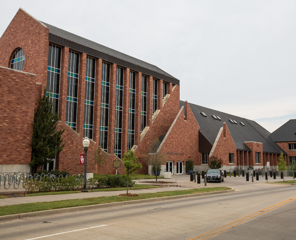 A large red brick building with modern, angular design features tall windows. A street with light traffic and a parked