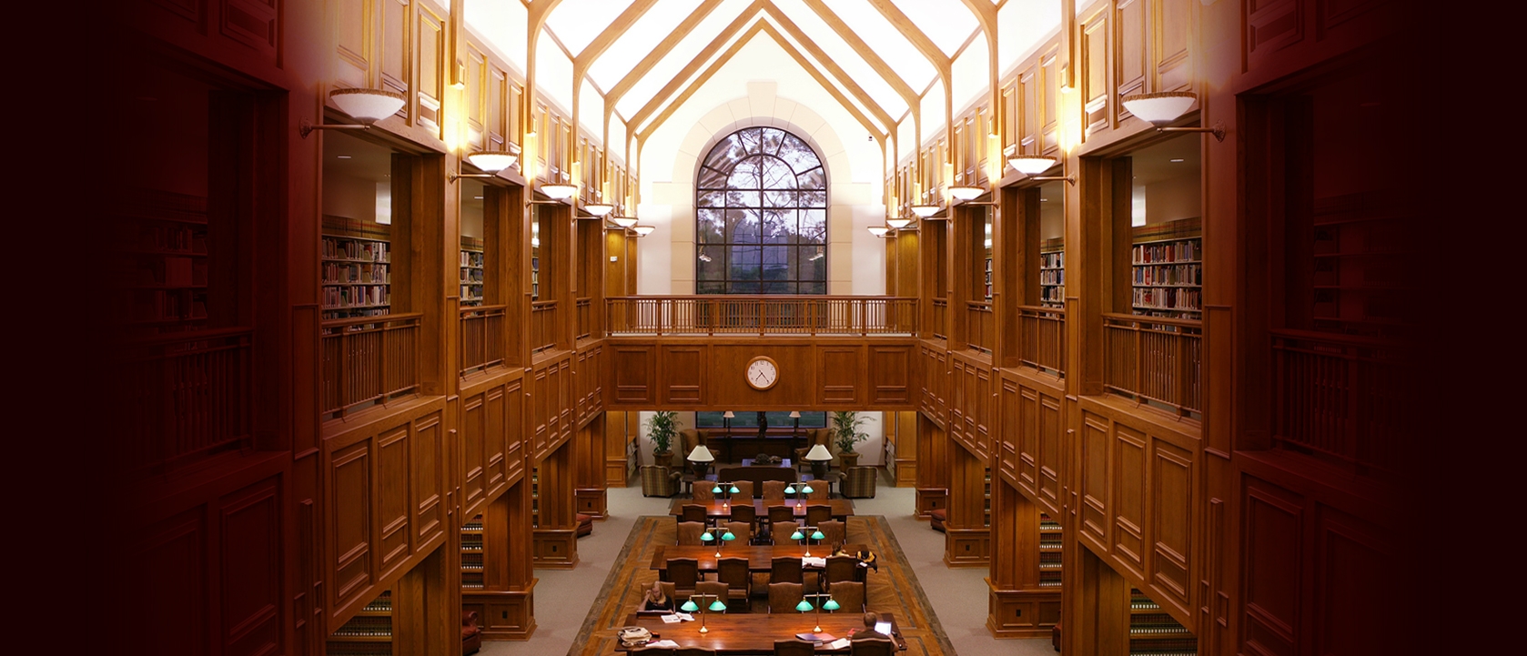 A spacious library interior with wooden shelves, a high ceiling, and a large arched window. Tables have green lamps. Warm lighting creates a cozy and studious atmosphere.