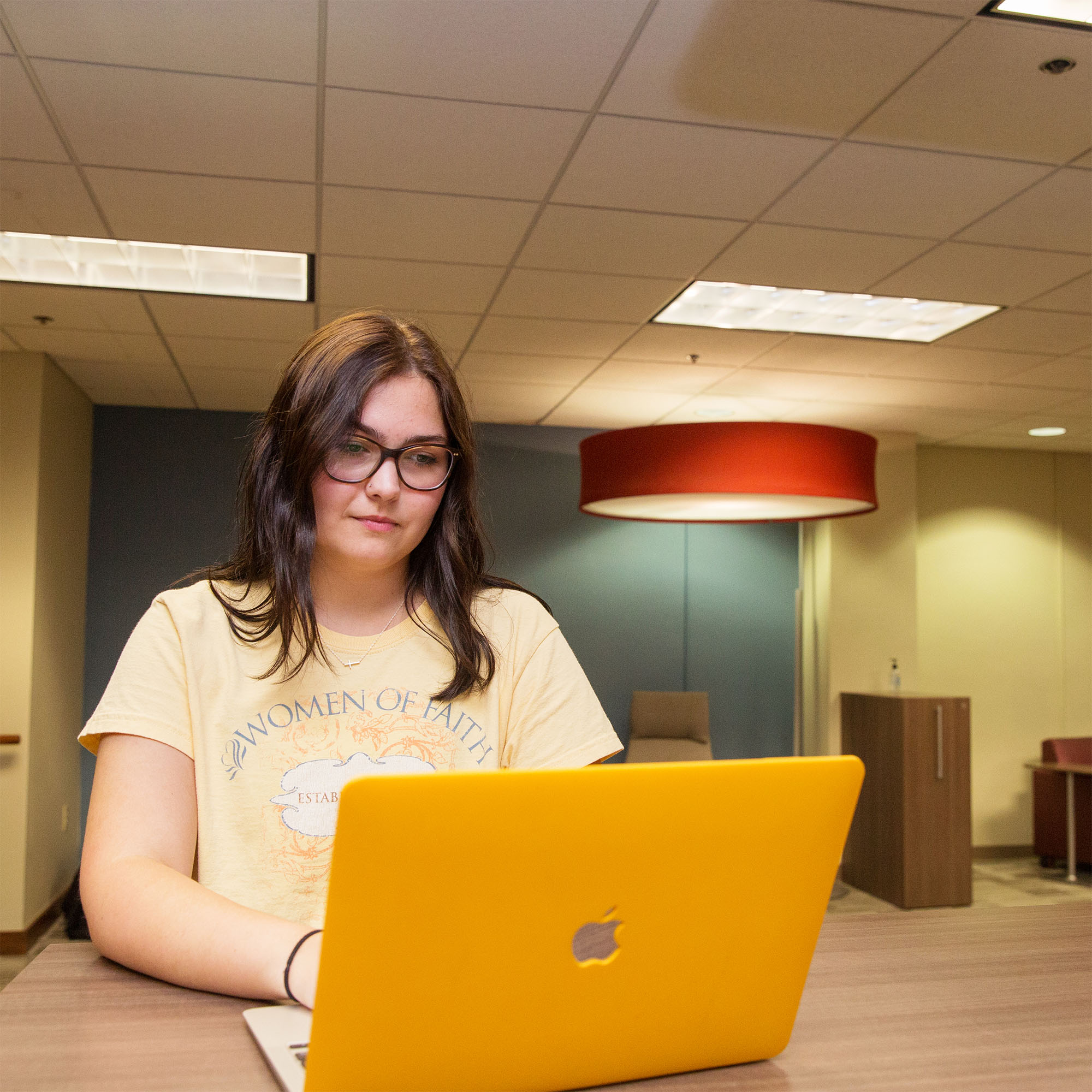 A female student sits at a table in a quiet room, working on her laptop and focusing on the screen as she types.