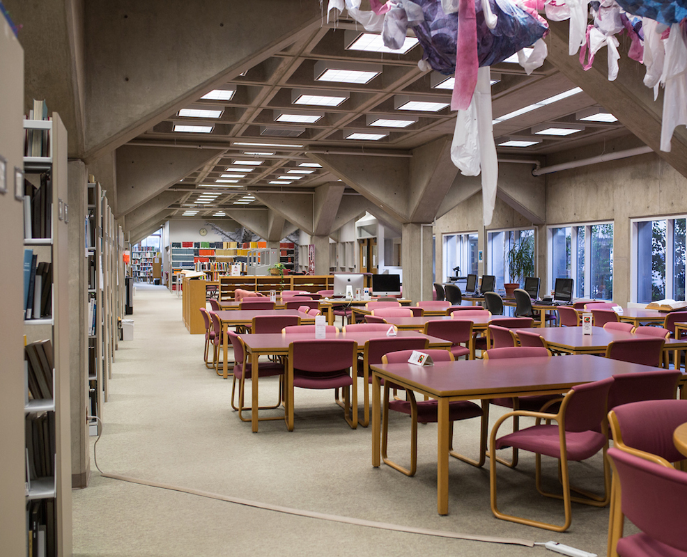 The Fine Arts Library is a spacious interior with modern architecture, featuring rows of wooden tables and pink chairs. Bookshelves line the walls, and colorful fabric art hangs from the ceiling.