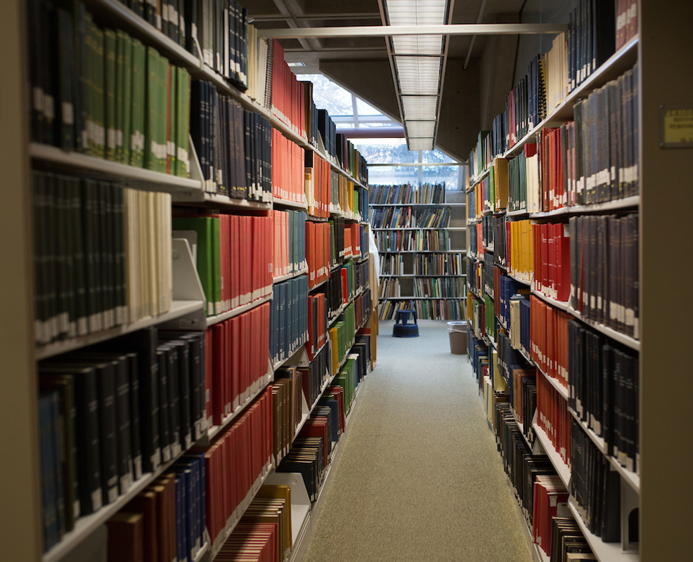 Rows of colorful books line a narrow library aisle, with natural light streaming through a window at the end. The space feels quiet and scholarly.