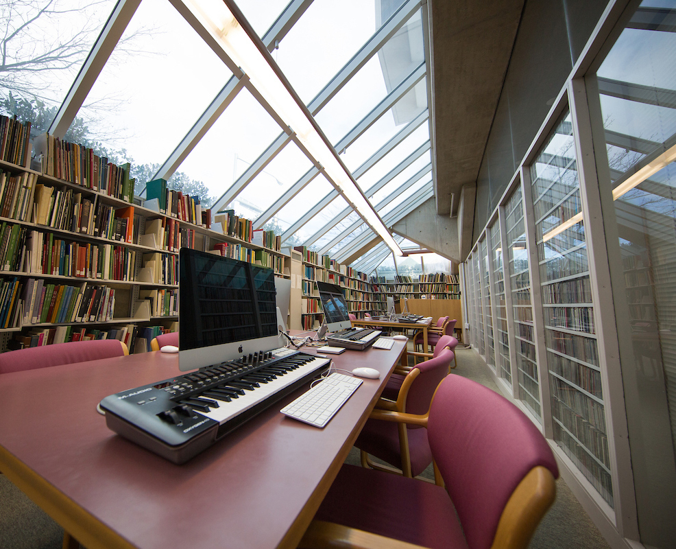 A modern room inside the Fine Arts Library features shelves filled with books, a long table, dark pink chairs, and Mac computer stations with MIDI keyboards, under a slanted glass ceiling with natural light coming in.