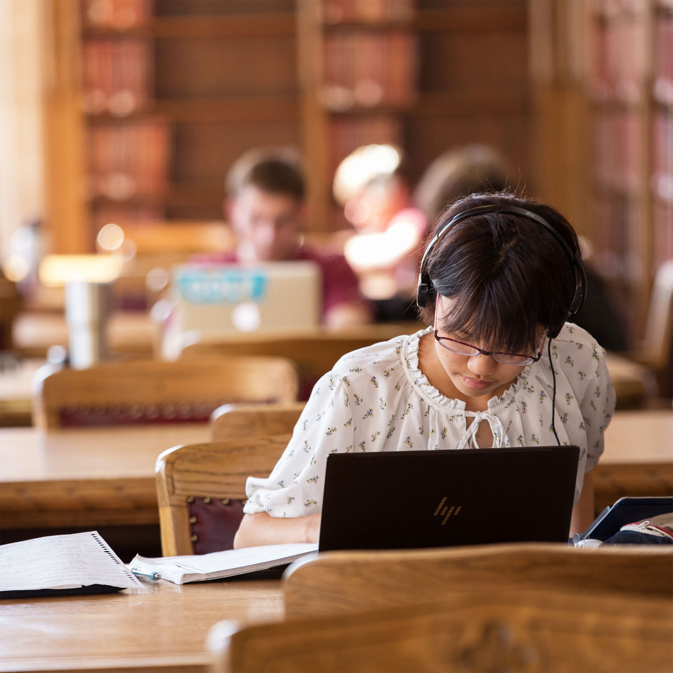 A person with headphones and glasses sits at a table focusing on a laptop in a library, surrounded by wooden tables and blurred background figures, creating a studious atmosphere.