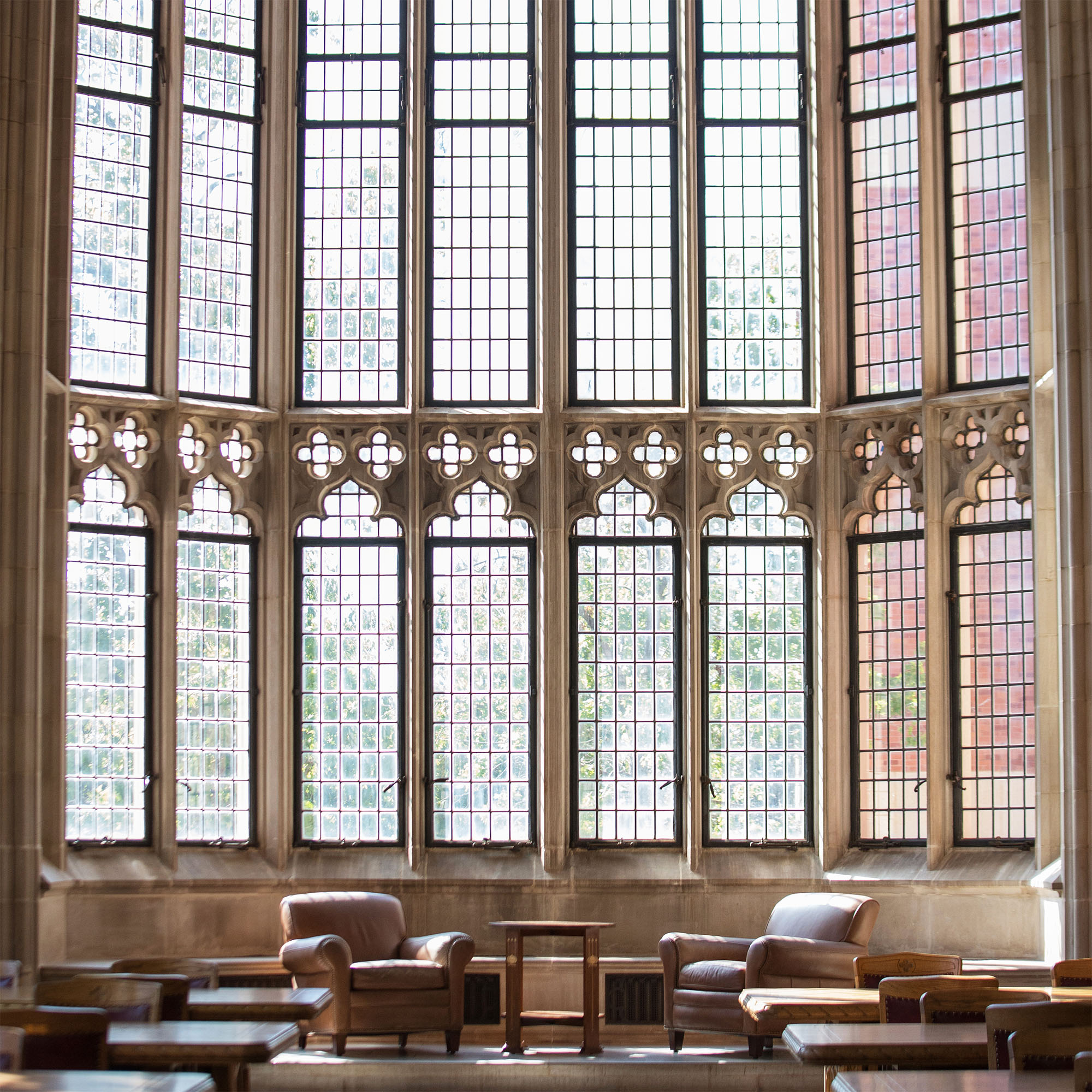 A spacious library reading hall with tall, ornate glass windows, two cozy leather chairs with small round wooden table between them and rows of long wooden tables with chairs to sit and study at.