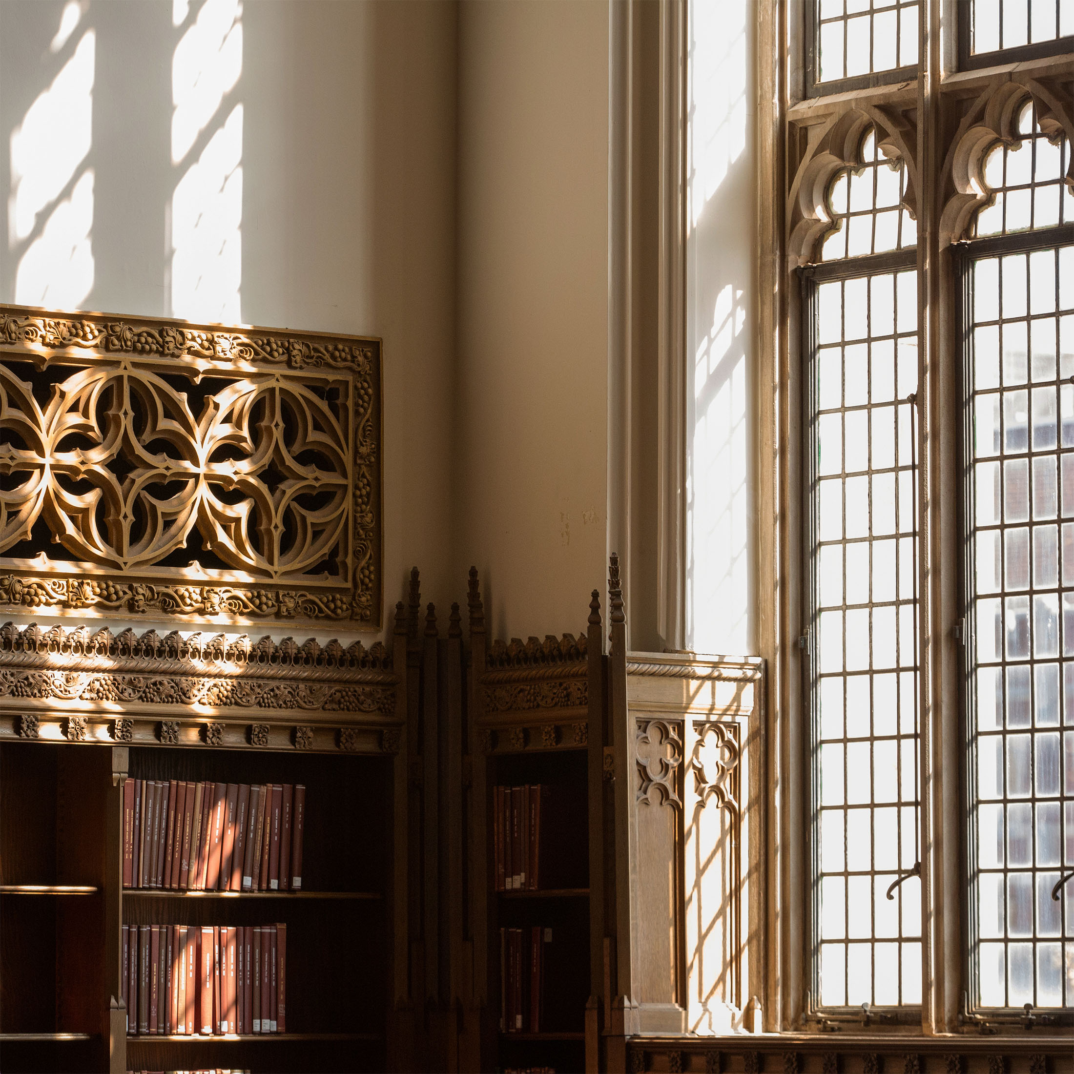 Sunlight streams through tall, ornate windows of the historic Helmerich Great Reading Room, casting intricate shadows on wood carvings and illuminating shelves of books.