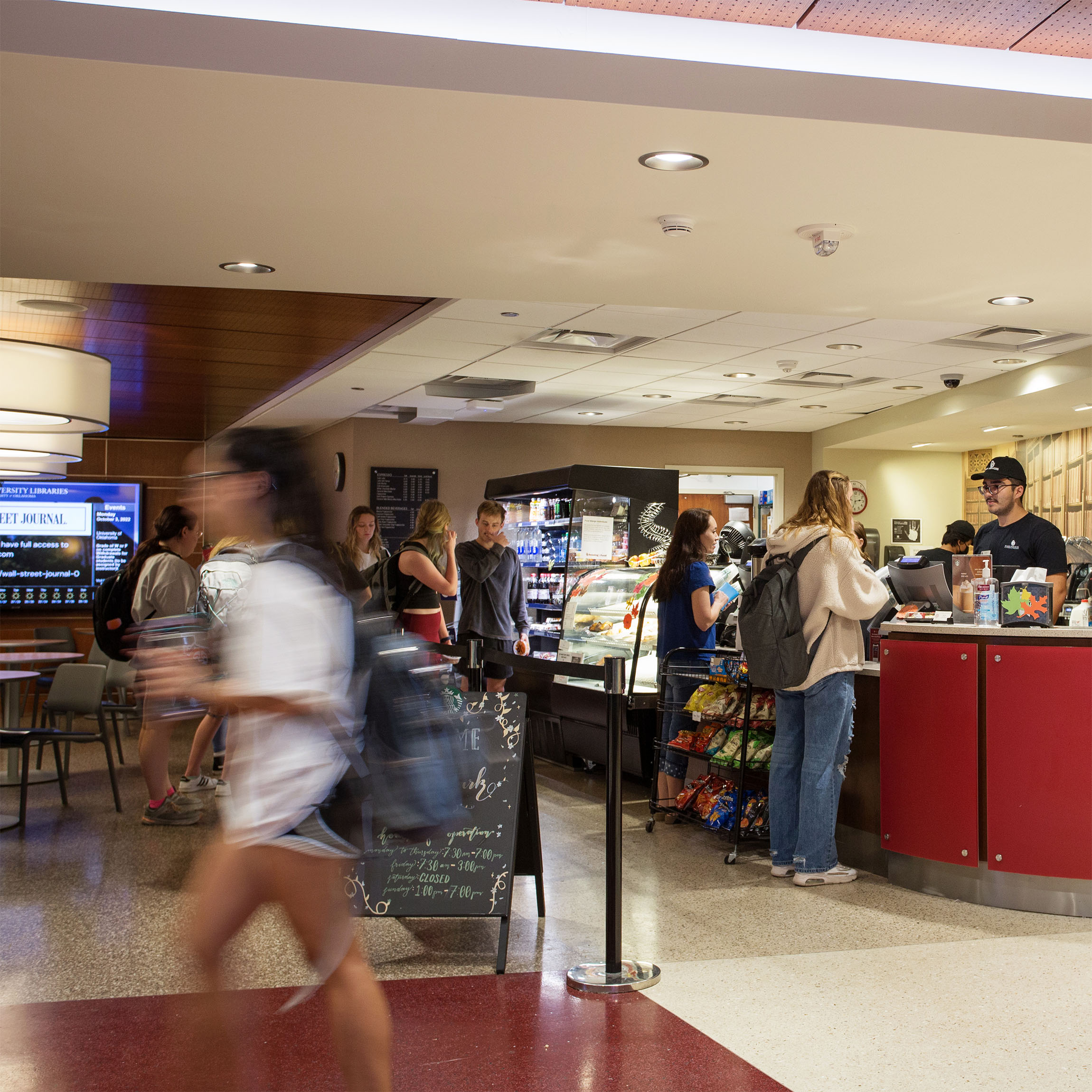 Busy café with a modern interior. People stand in line, interacting with a cashier at the counter. Customers and students are seen with backpacks.
