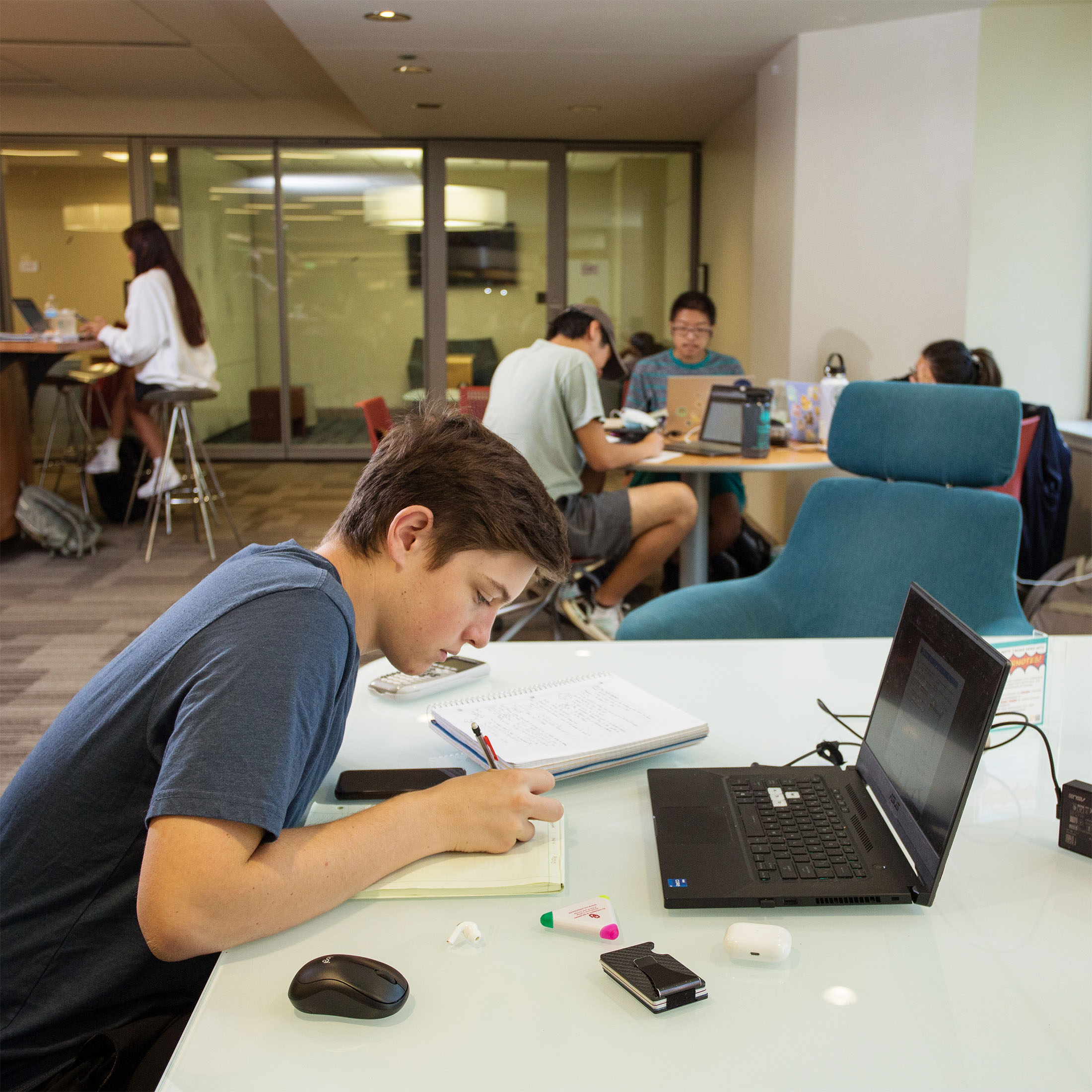 A focused young person writes in a notebook at a desk with a laptop, surrounded by study materials. In the background, students collaborate in a study room.