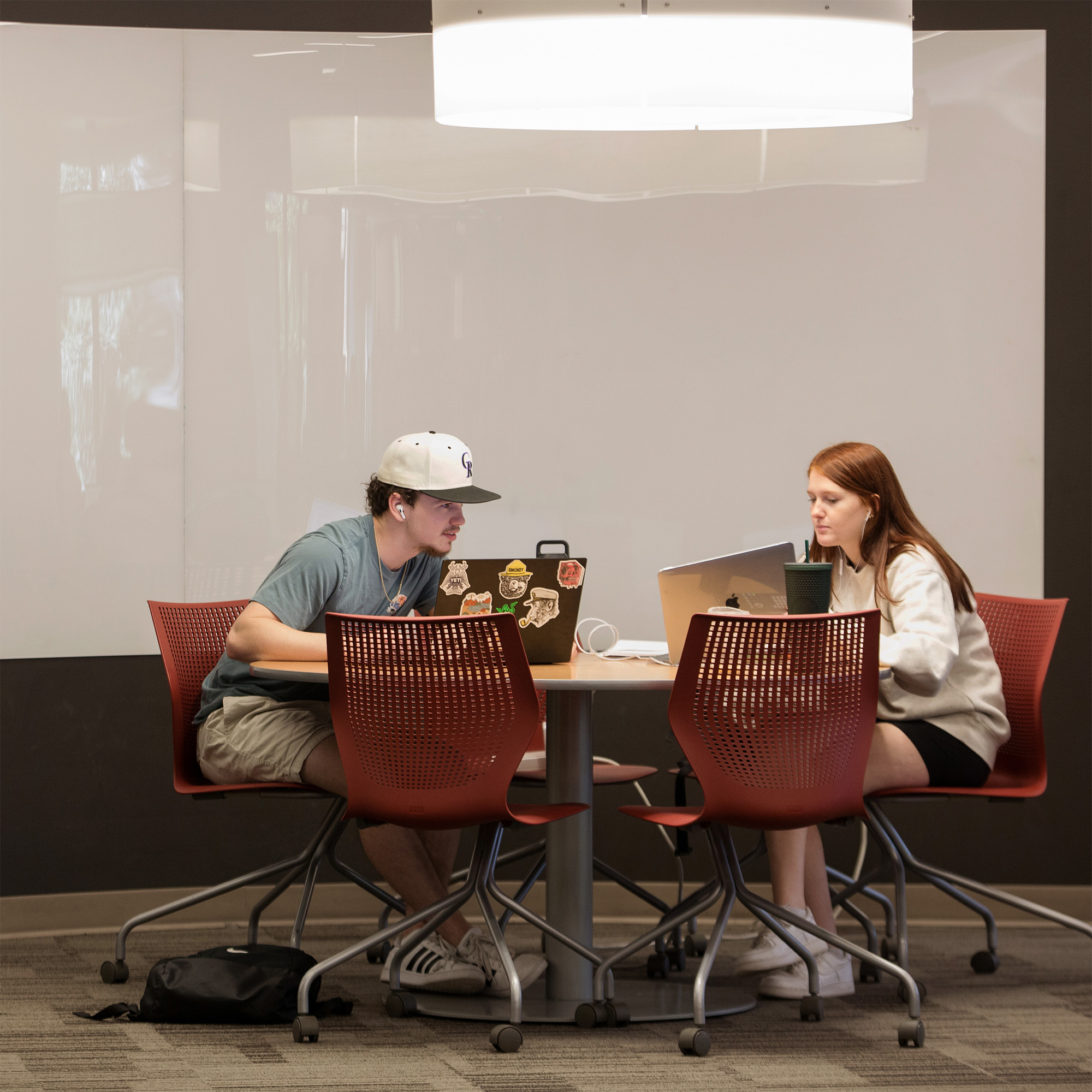 Two students sit across from each other at a circular table with laptops, focused and engaged. Red chairs, soft lighting, and a blank whiteboard set the study room scene.