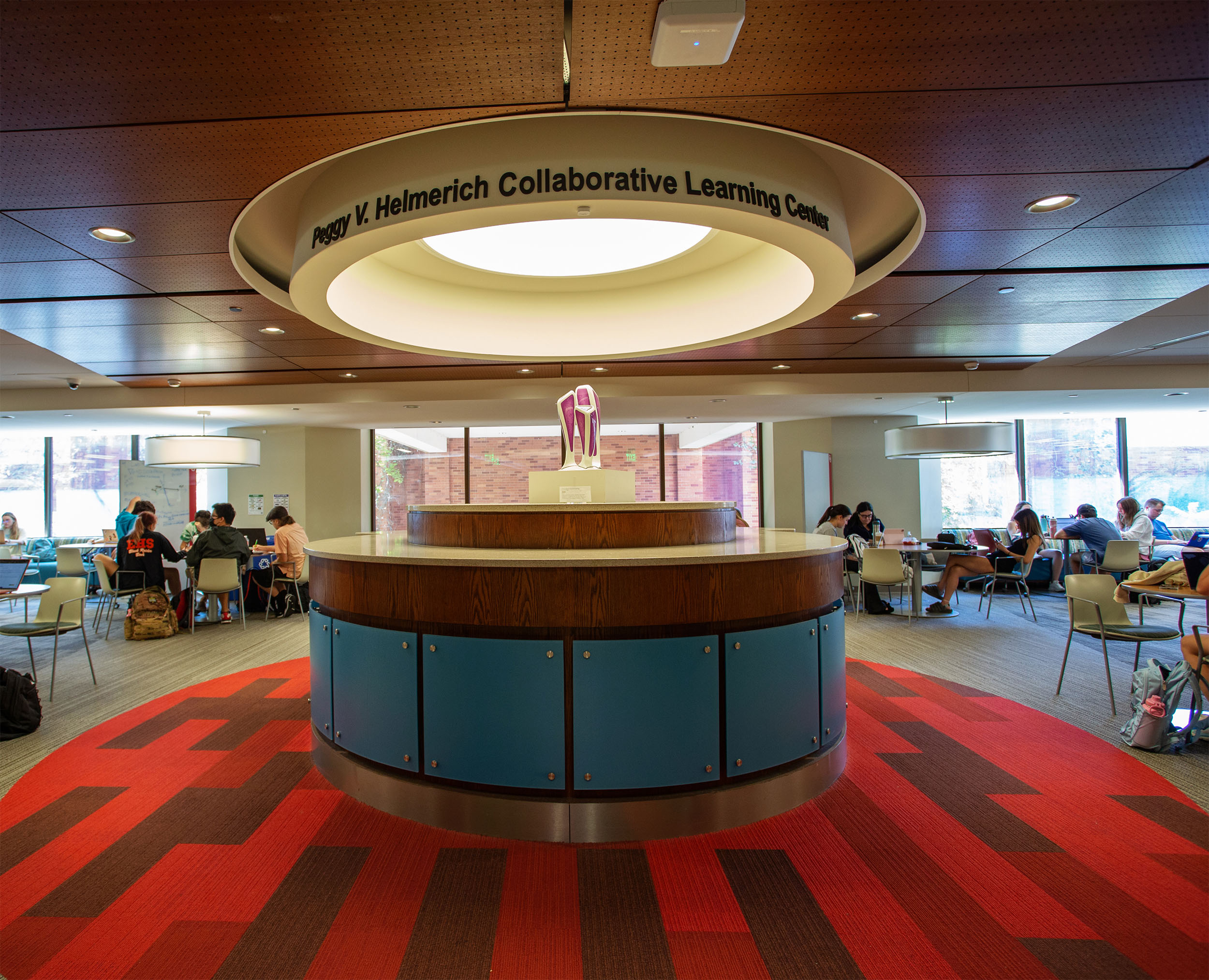 A modern study area in the Peggy V. Helmerich Collaborative Learning Center with red carpet, circular wooden desk, and students working at tables, creating a focused atmosphere.