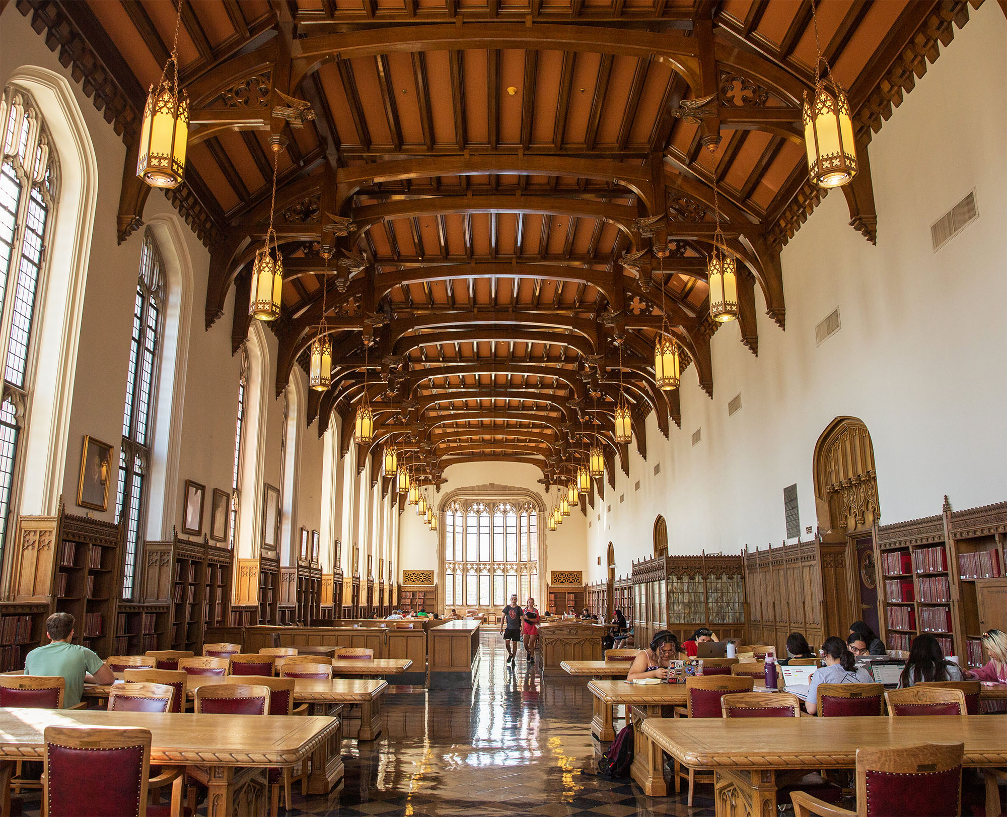 Grand library hall with vaulted wooden ceiling, large windows, and ornate chandeliers. Students sit at long wooden tables, studying. Warm, academic atmosphere.