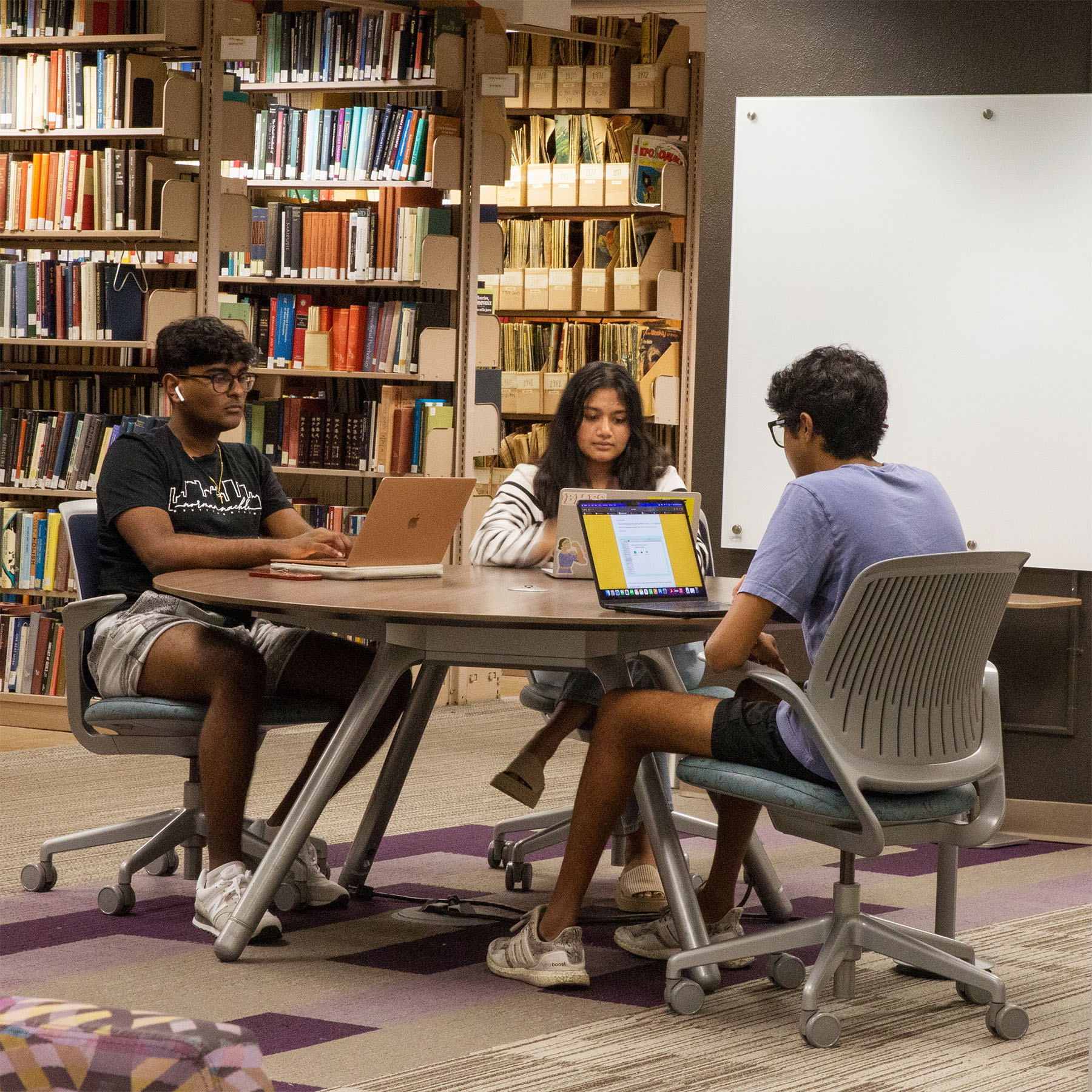 Three students sit at a round table with laptops in a library, surrounded by bookshelves. They appear focused and engaged in study.