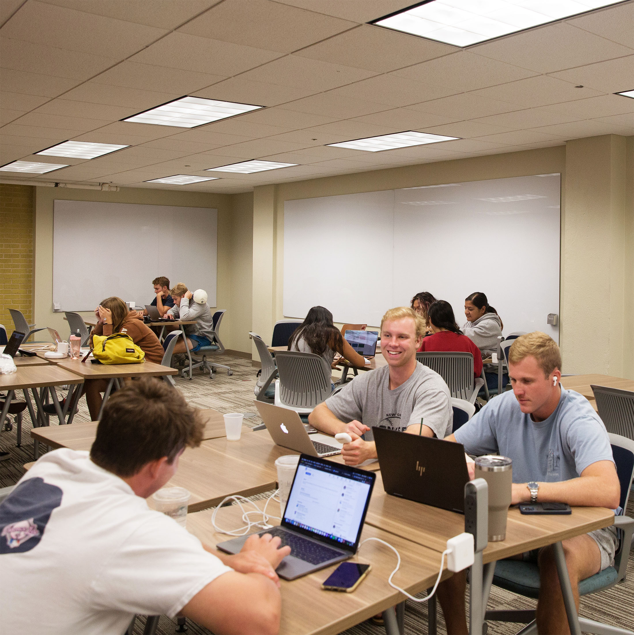 A group of students in a brightly lit classroom, seated at tables with laptops. They are focused and engaged, creating a collaborative and productive atmosphere. Large white boards on the walls in the background.