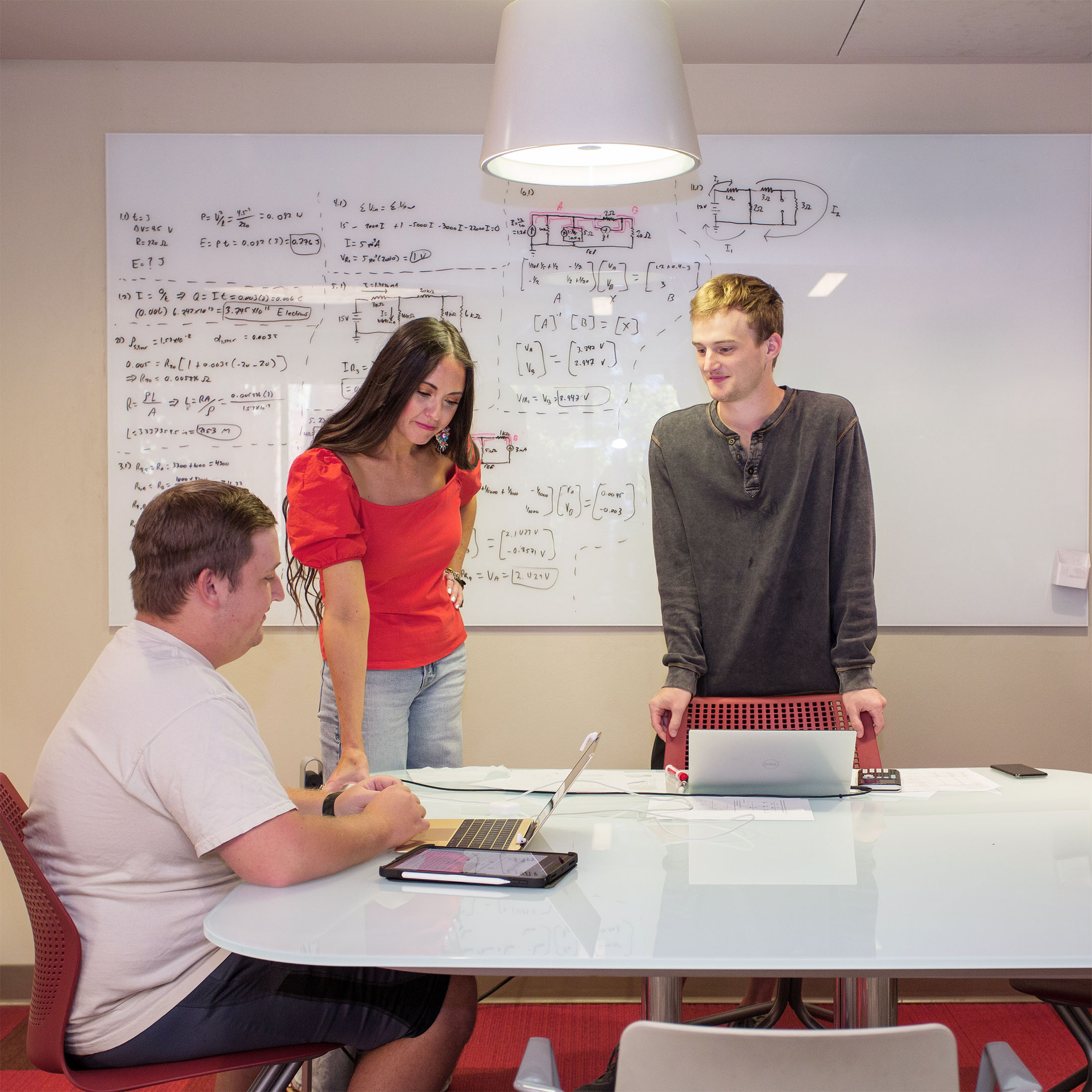 Three people collaborate in a modern library work room setting. They are gathered around a table with laptops, engaged in discussion. A whiteboard with diagrams is in the background.