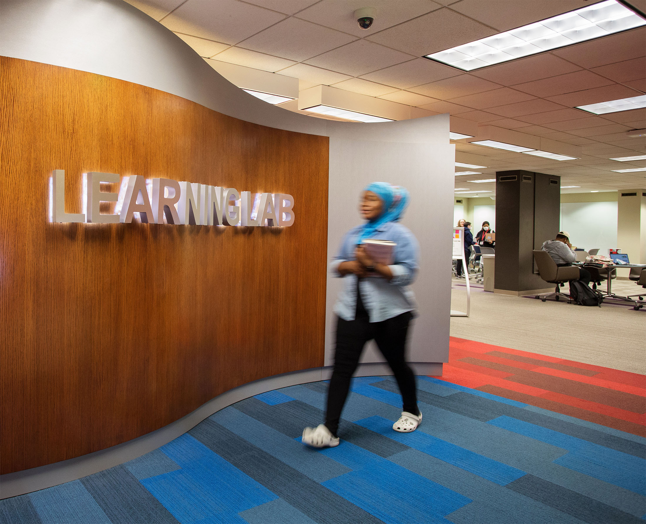 A person walks past a "Learning Lab" sign on a wooden wall in a modern, brightly lit study area, wearing a blue hijab and carrying books.