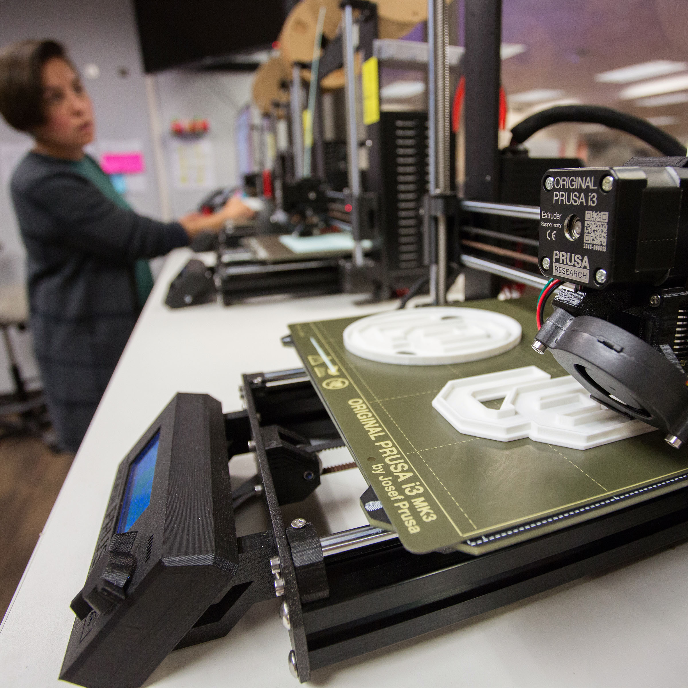 3D printers are set up in a row on a workbench. In the forefront is a close-up of a 3D printer producing white OU plastic logos. A person is working in the background, focusing on the task, creating a tech-savvy atmosphere.