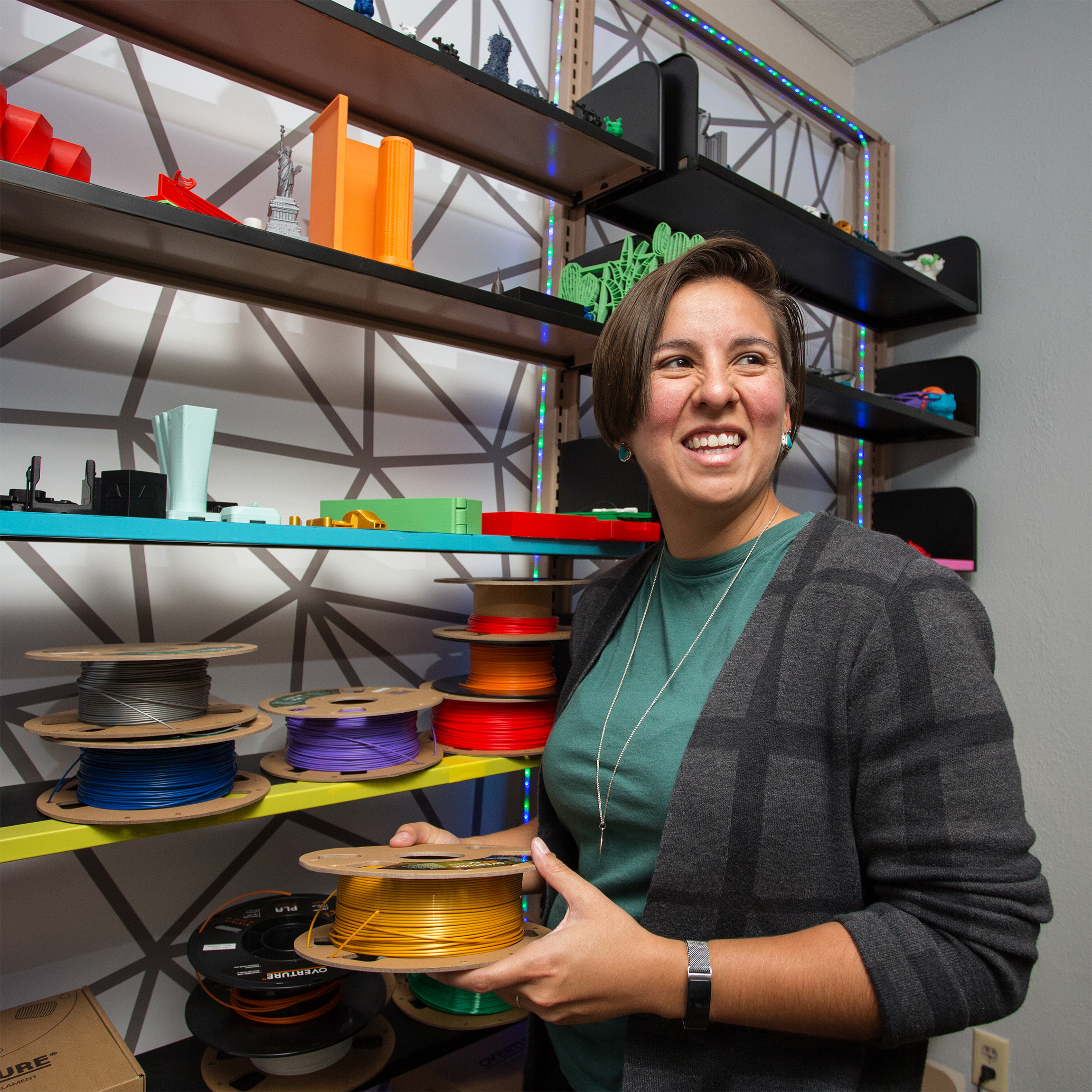 A smiling OU Libraries employee stands beside shelves with colorful 3D printed models and filament spools. The atmosphere is creative and vibrant.