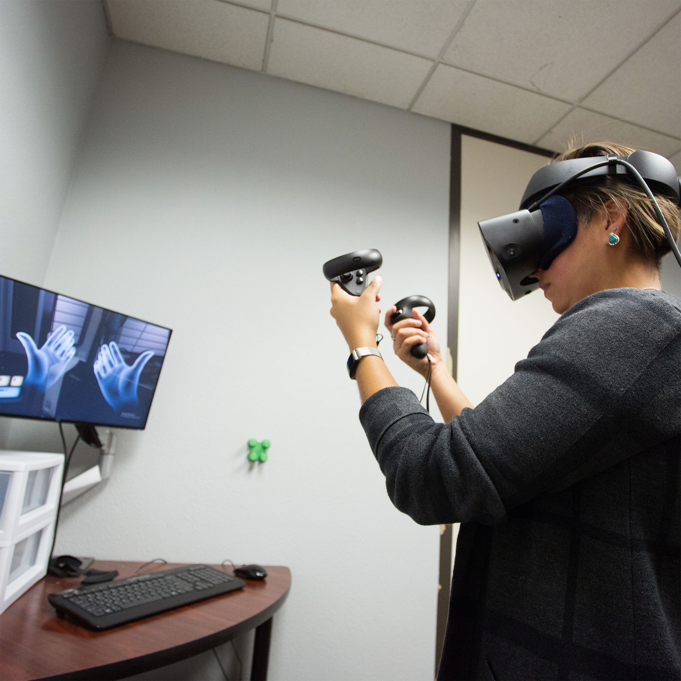 A person wearing a VR headset and holding controllers in both hands engages with virtual reality in a room. A monitor shows virtual hands, suggesting immersion.
