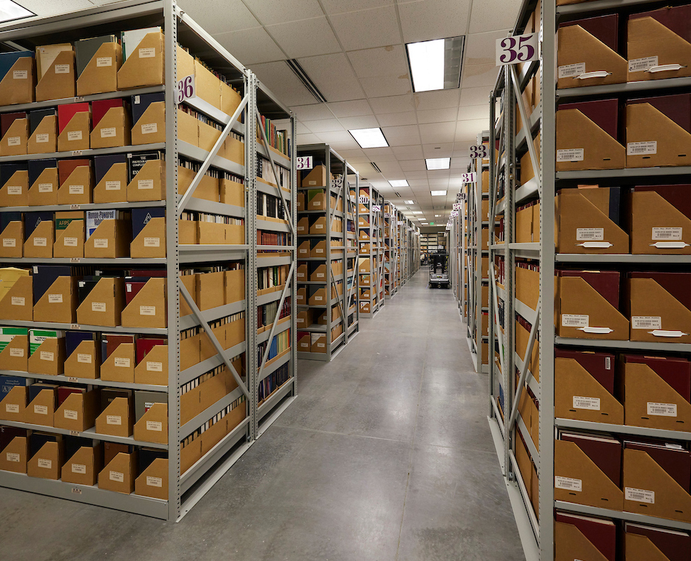 A well-lit archive with tall metal shelves is filled with labeled brown boxes, creating a sense of organization and vastness; a narrow aisle stretches into the distance.