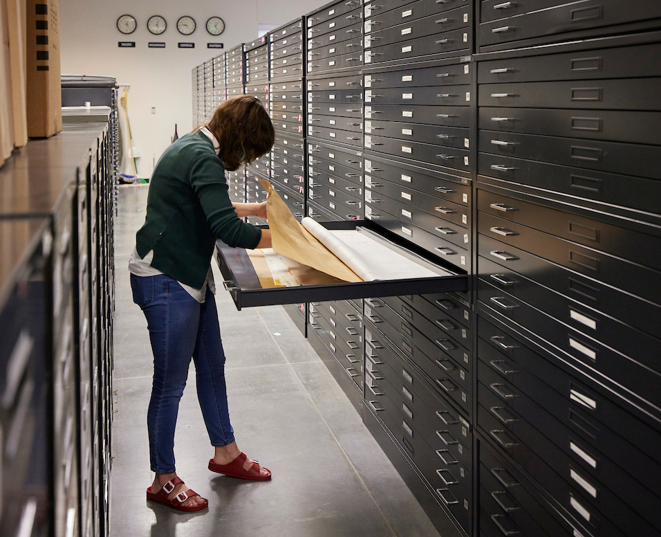 A person in a green sweater and jeans examines documents in a long, flat drawer in an archive room, surrounded by tall, metal cabinets and clocks on the back wall showing different time zones.