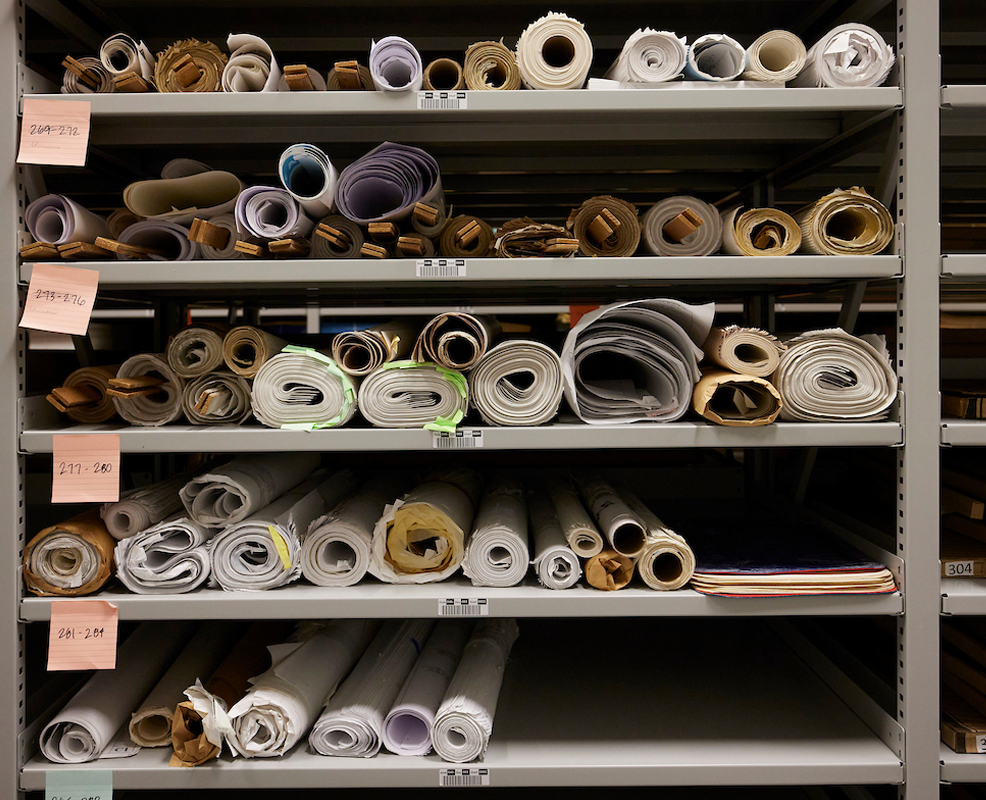 Metal shelves are filled with neatly organized, rolled-up documents such as architectural blueprints. The rolls vary in size and color and are labeled with sticky notes.