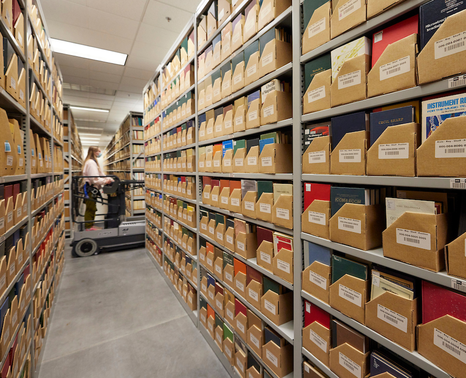 In the Library Service Center, rows of shelves are filled with neatly organized boxes containing books and library materials, creating a sense of order and focus. A library employee in the background stands on a cart that is used to retrieve items on high shelves.